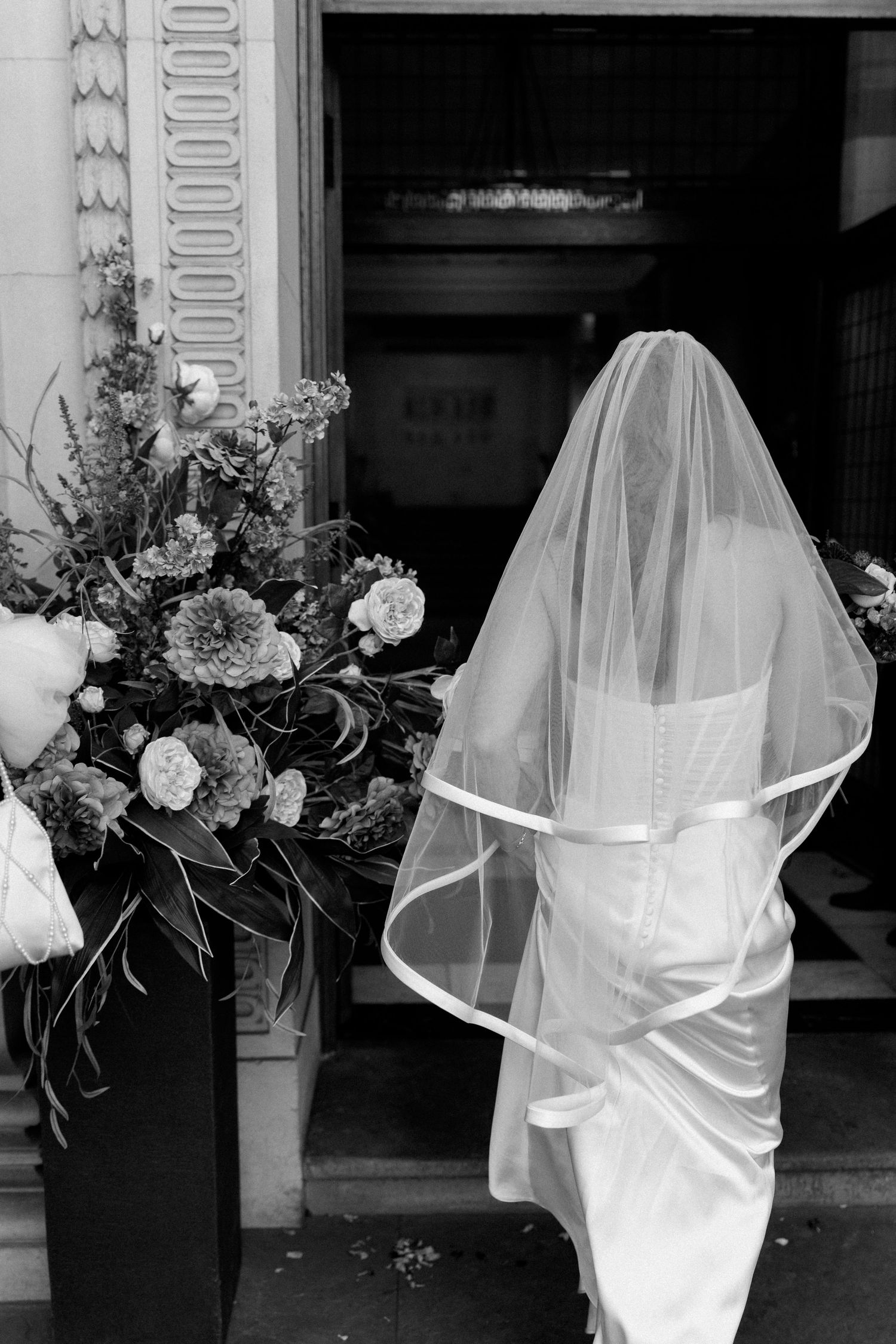Black and white photo of a bride in a white gown and veil entering a church doorway beside a floral arrangement.