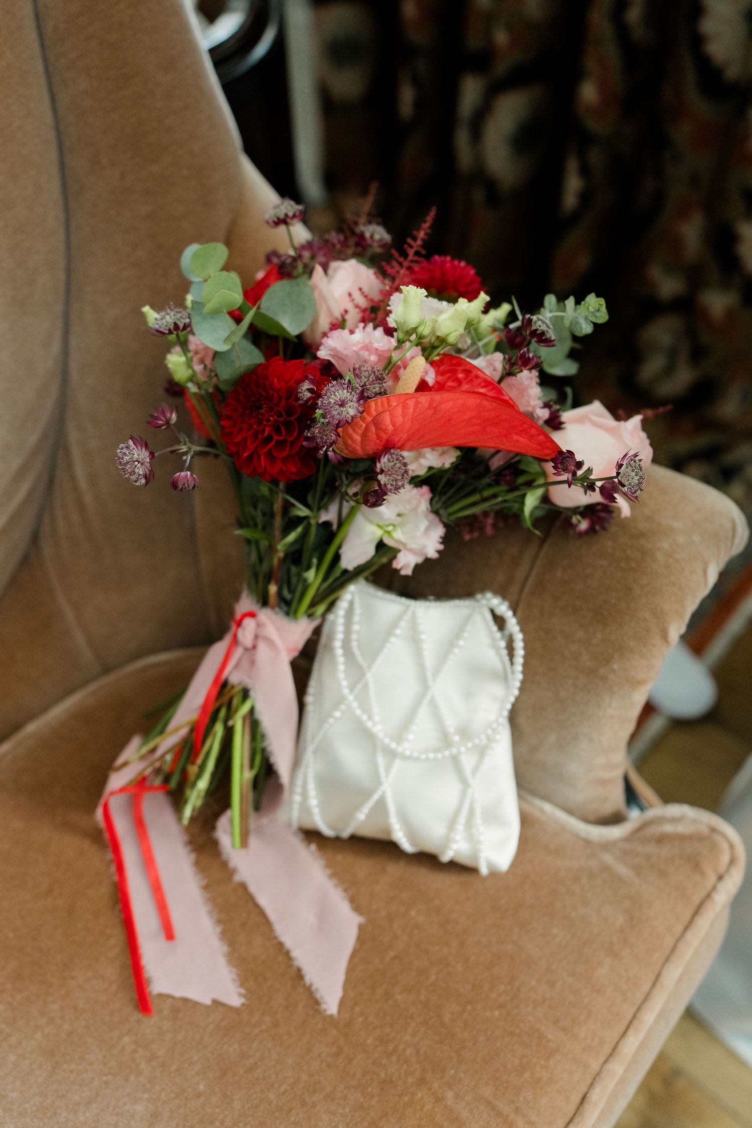 Red and pink bridal bouquet with eucalyptus beside a white pearl beaded purse on a beige velvet chair.