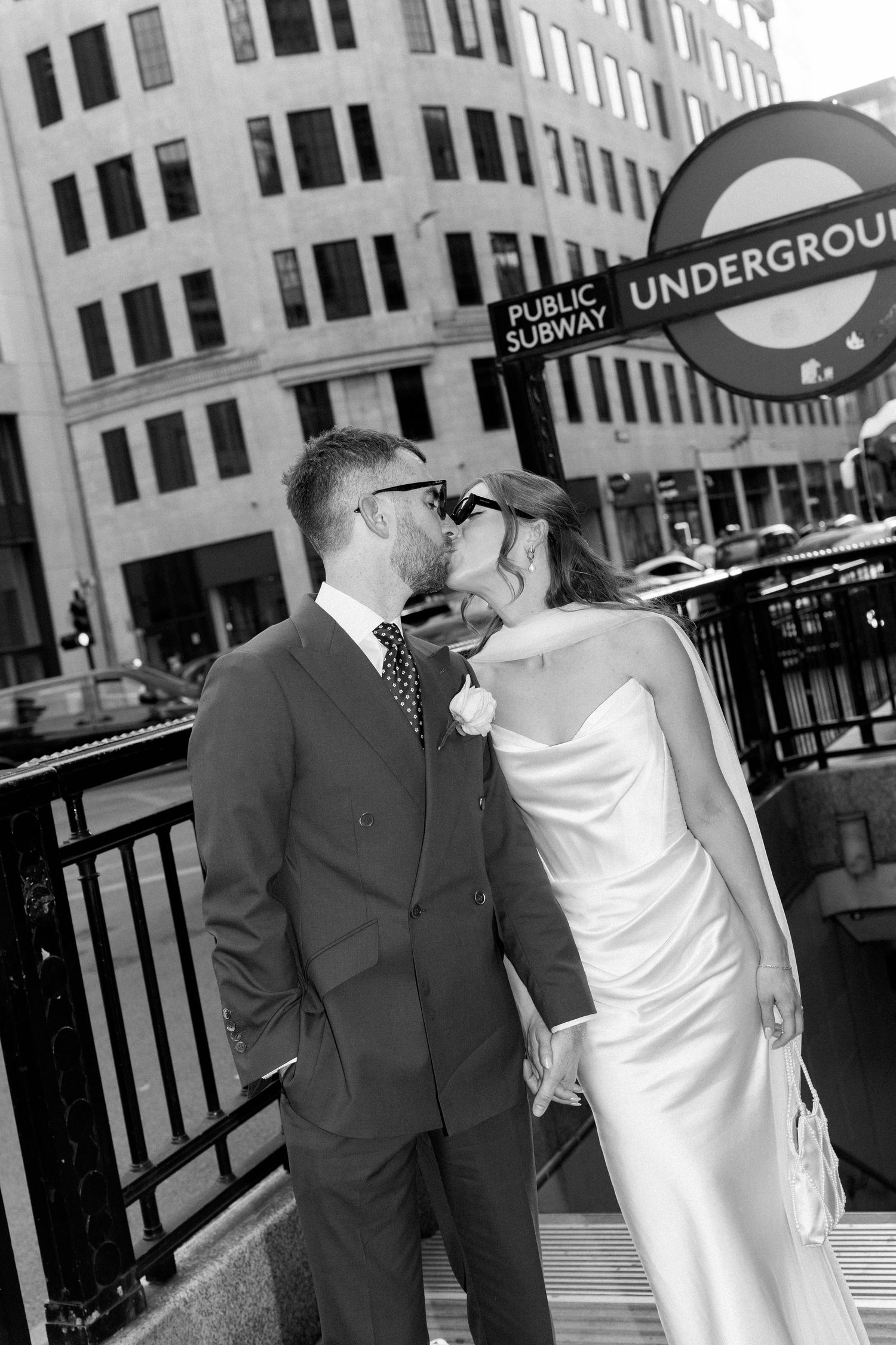 Bride and groom share a kiss beside iconic London Underground sign in black and white photo.