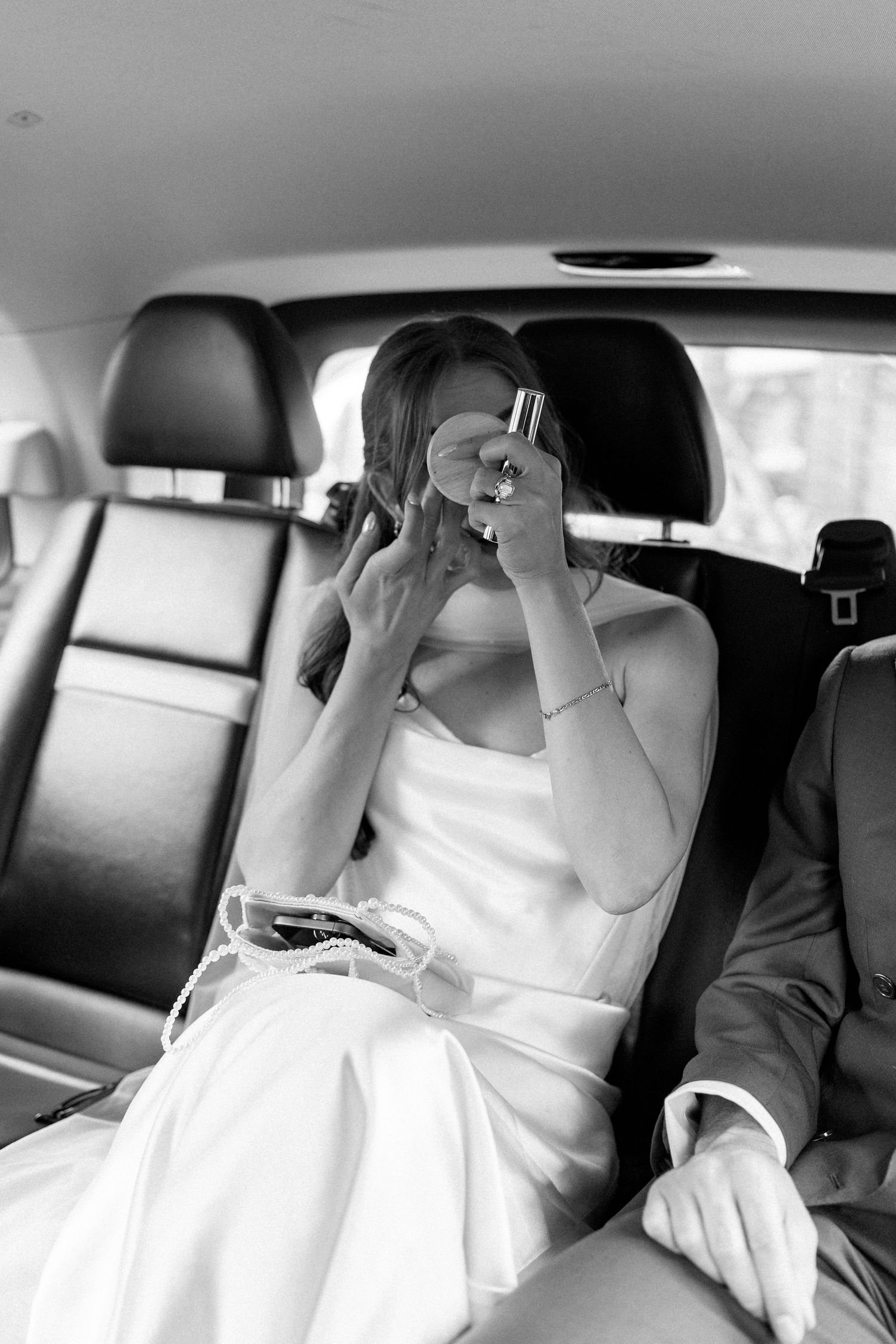Black and white photo of bride applying perfume in car backseat on wedding day.