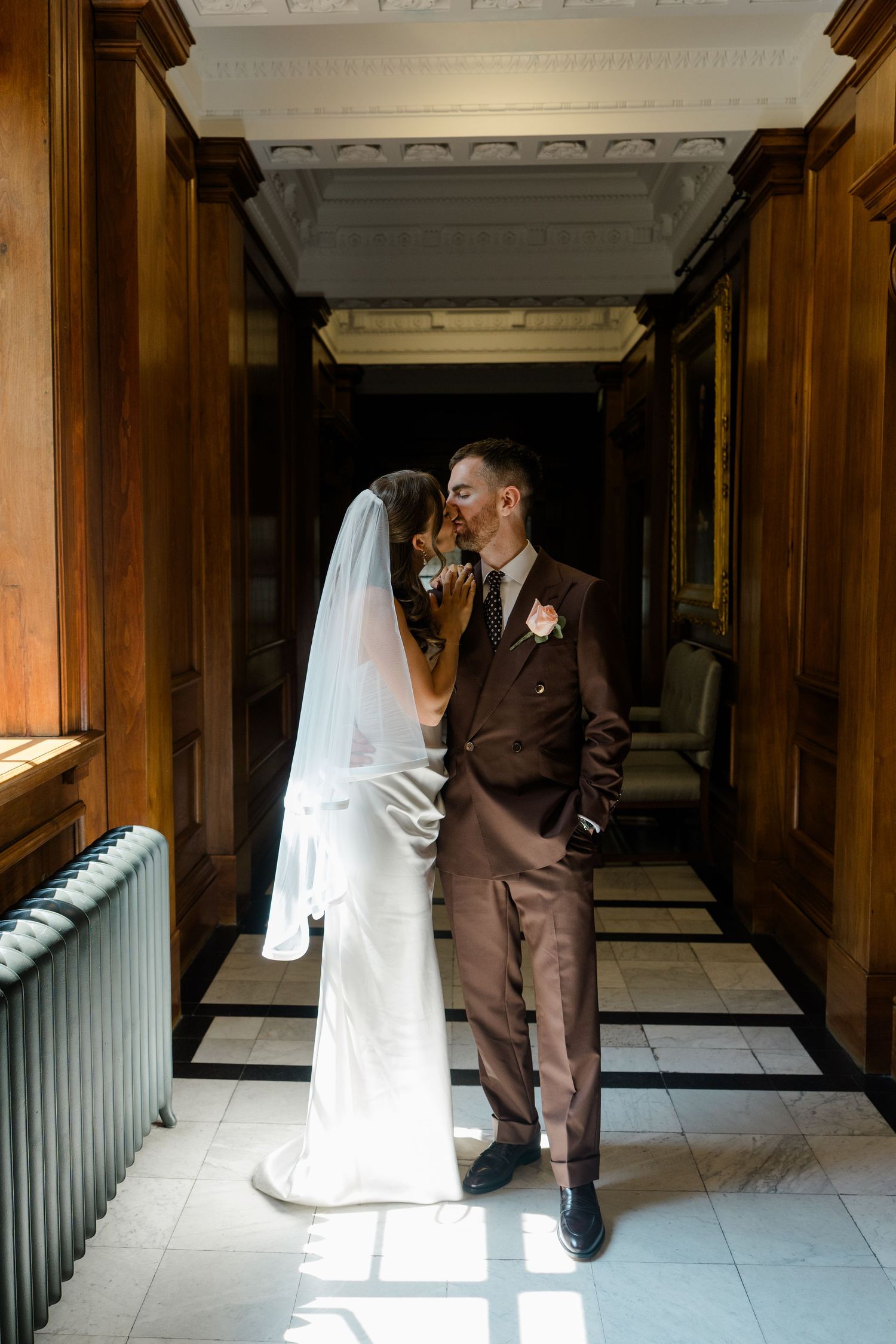 Bride and groom share a romantic kiss in a sunlit wood-paneled hallway on their wedding day.