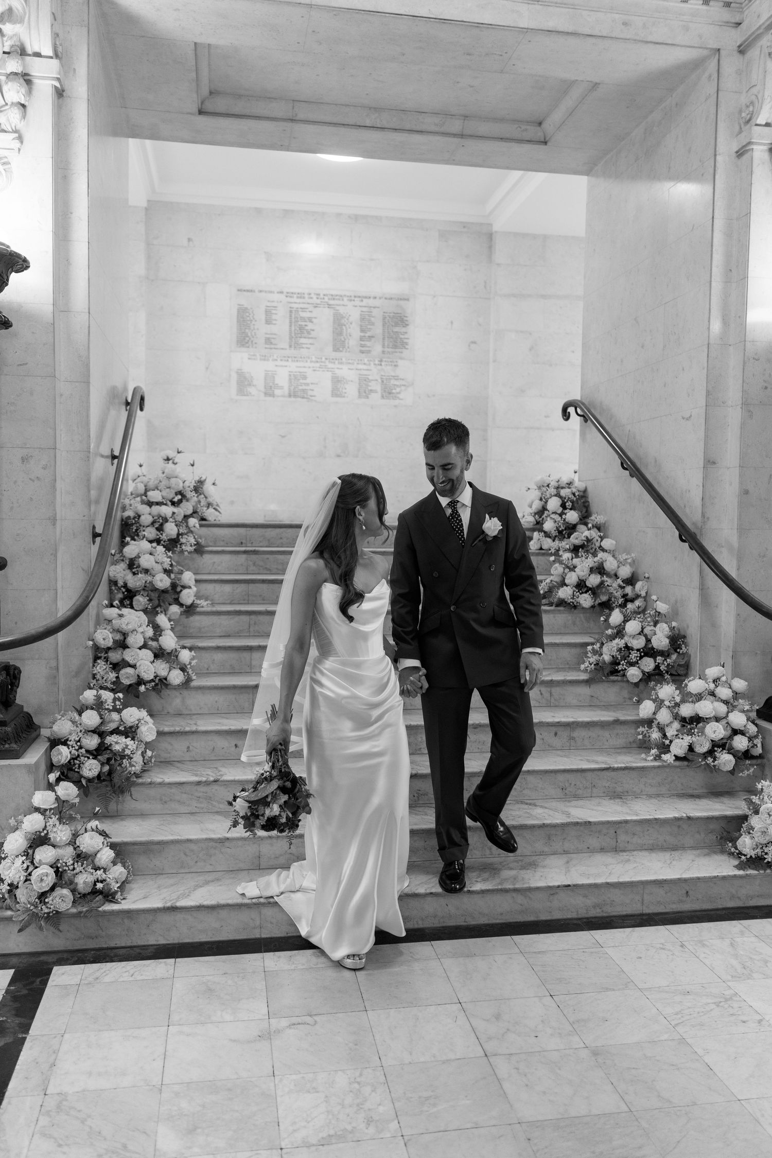Black and white photo of newlyweds descending floral-decorated stairs in elegant marble venue.