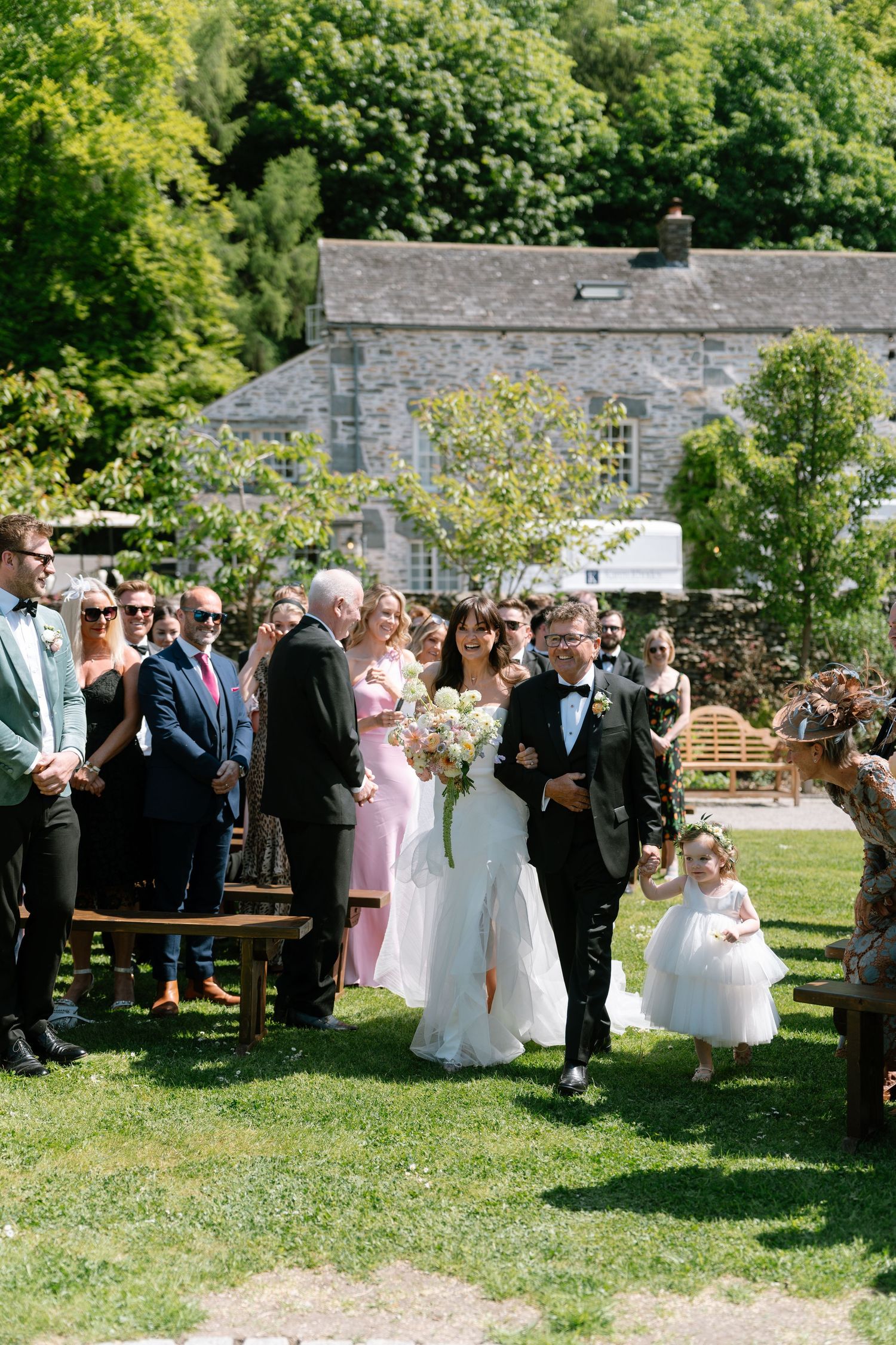 Wedding party and guests assembled on a grassy lawn near historic stone buildings.