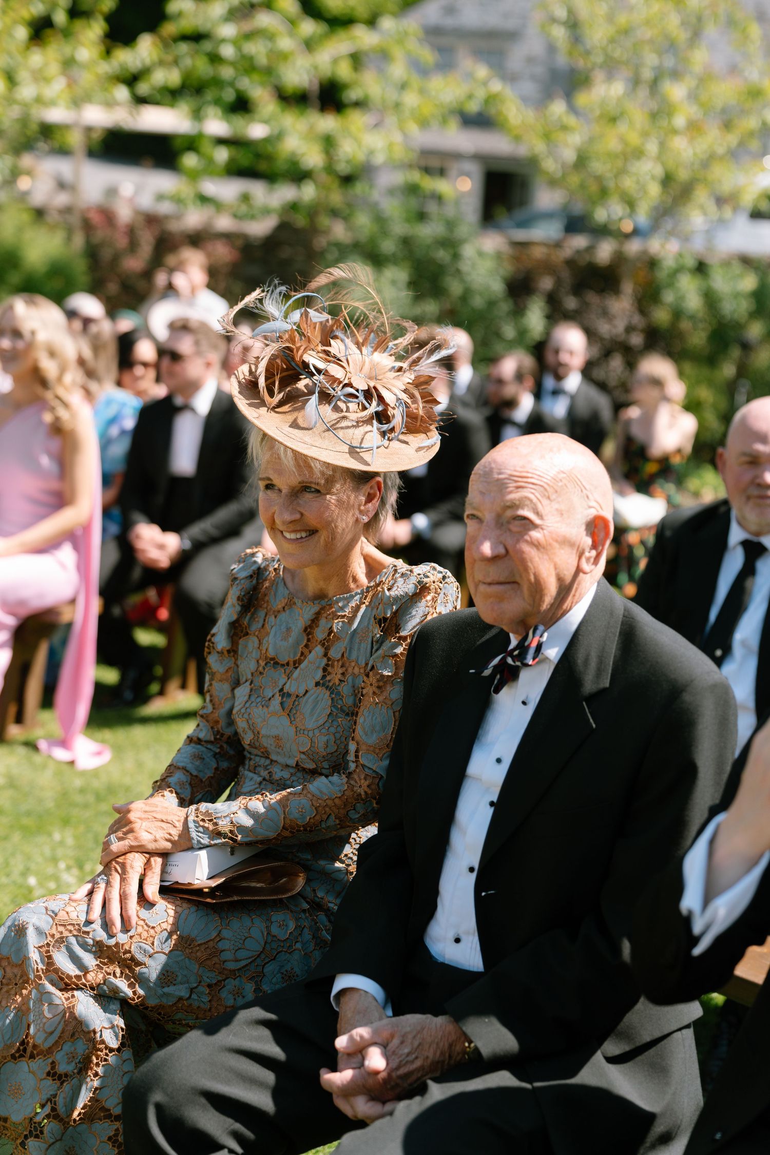 Elderly couple shares a joyful moment at an outdoor formal event with guests seated in the background.