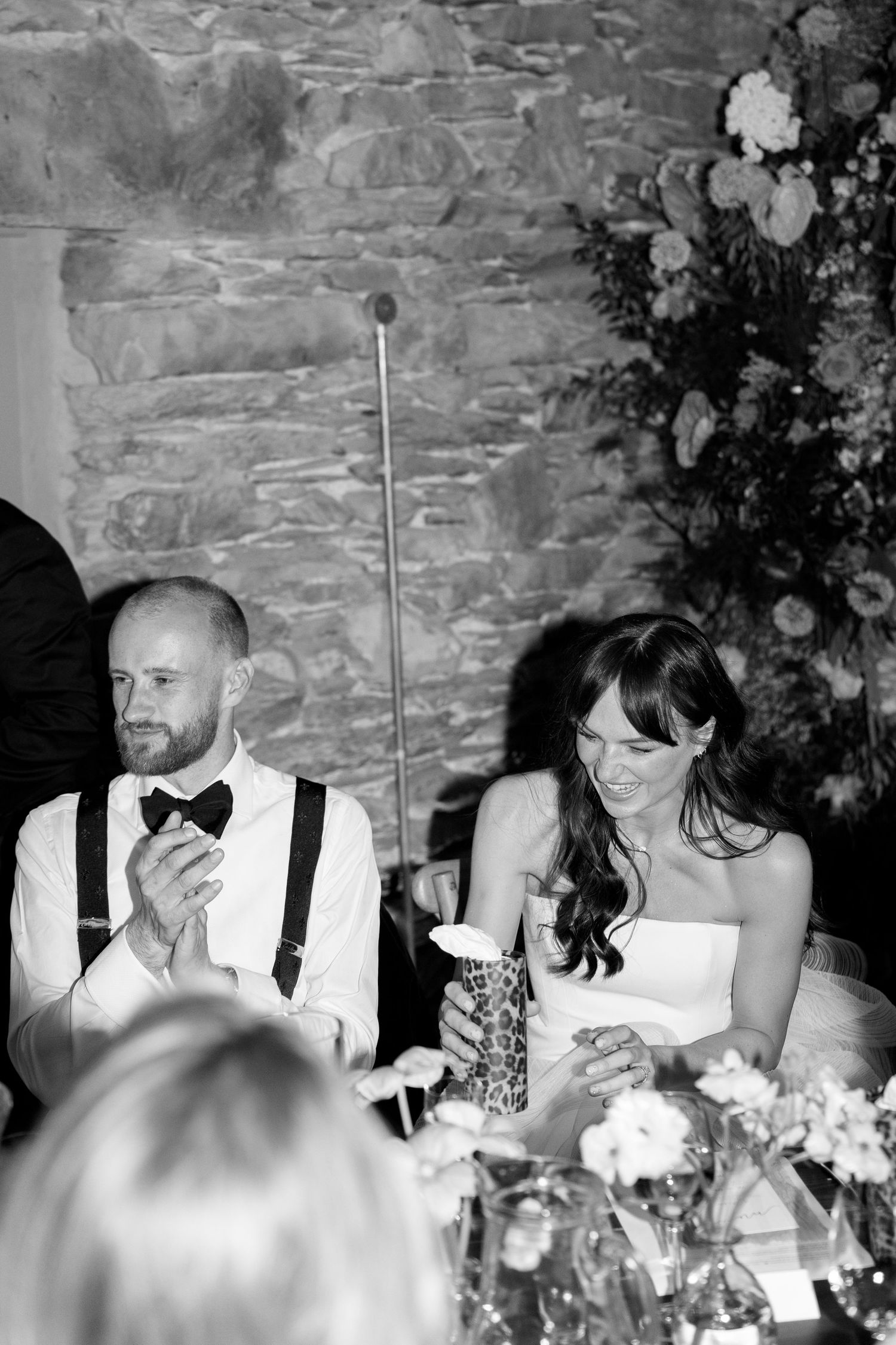 Wedding reception dinner table with guests in formal attire against rustic stone walls.