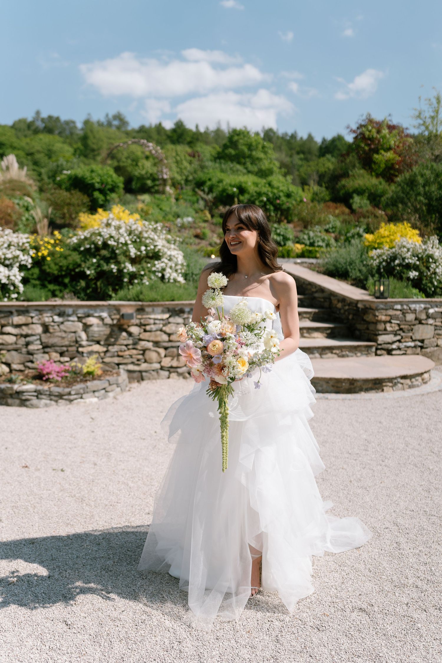 A bride in a flowing gown walks through a stone-walled garden carrying a bouquet.