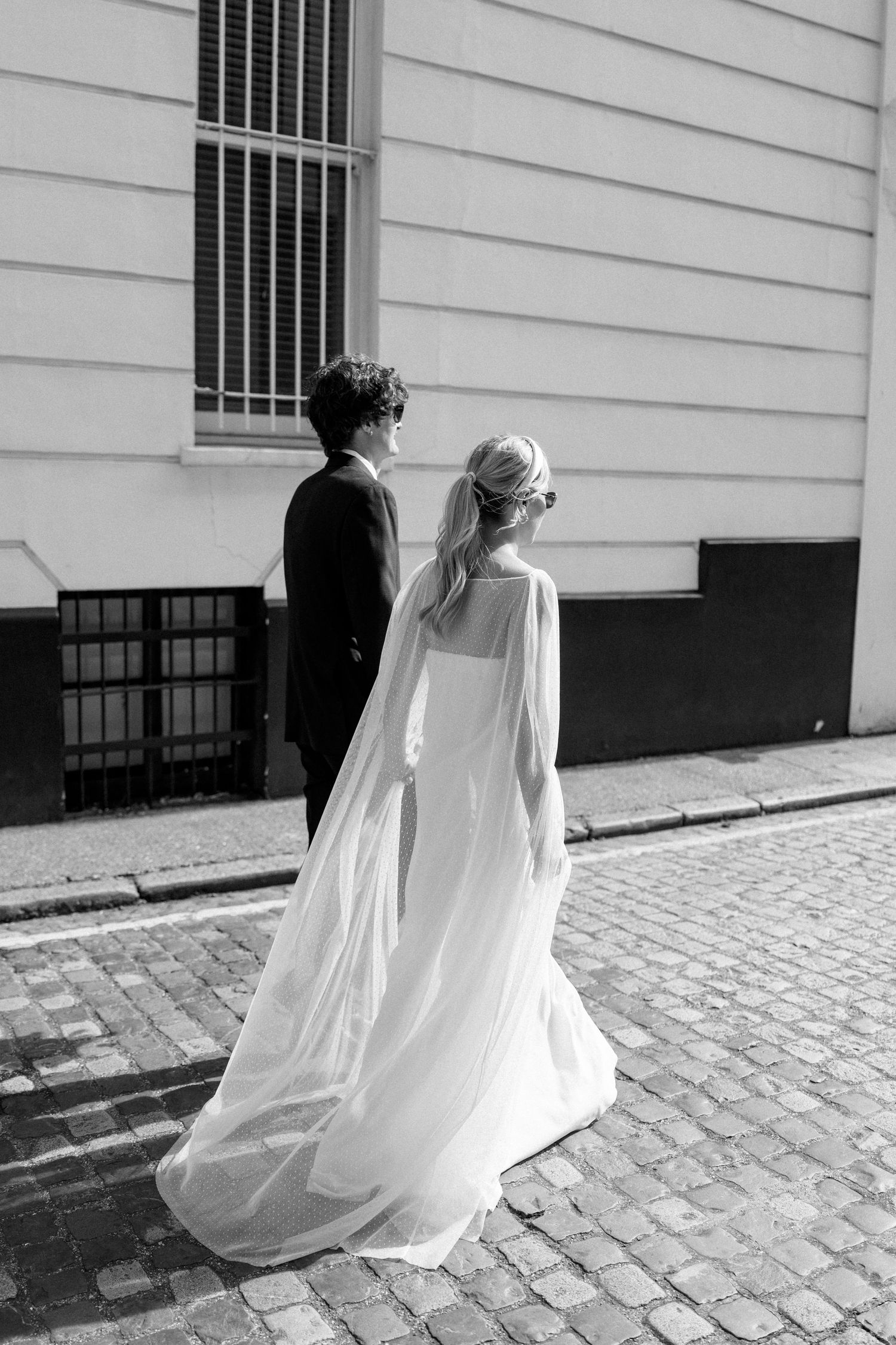 A flowing white wedding dress trails behind on cobblestone streets in dramatic black and white.