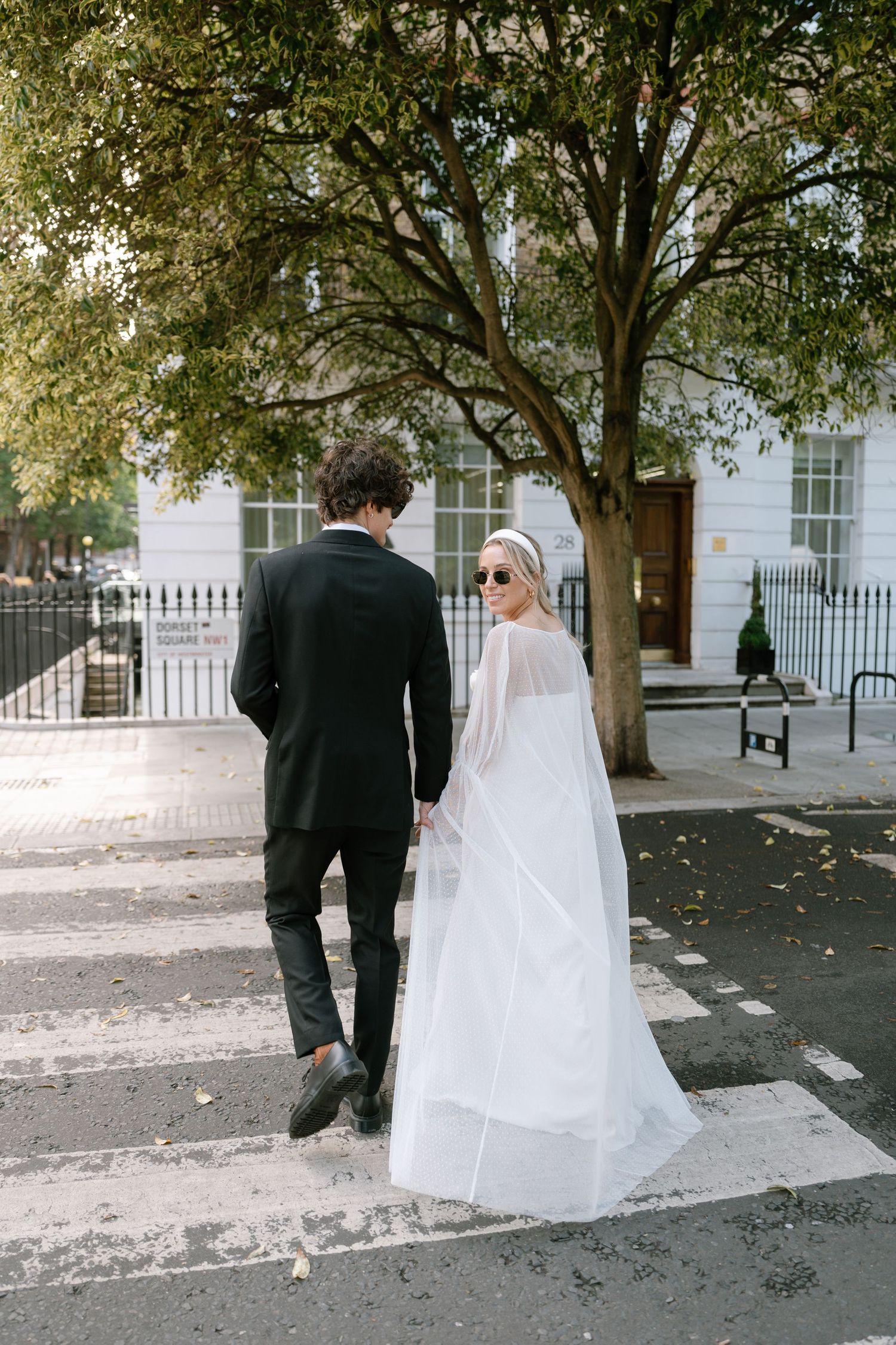 A couple walks down a tree-lined urban street with white buildings in the background.