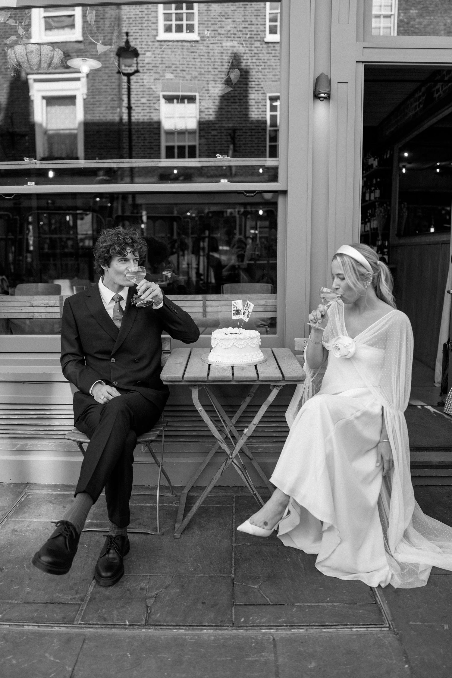 A couple shares a drink next to their wedding cake on an outdoor patio in black and white.