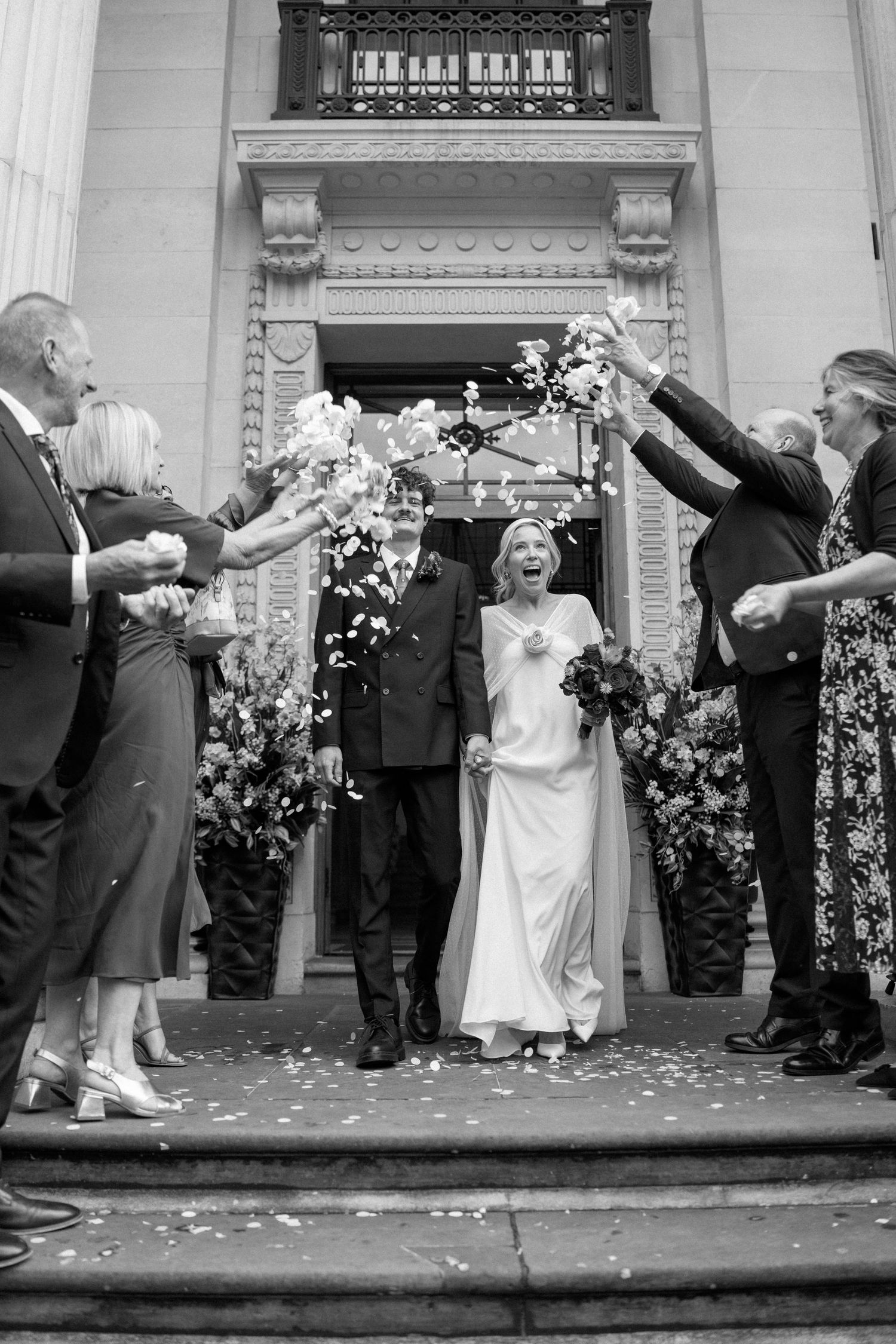 Wedding couple and guests celebrate with confetti outside a grand columned building entrance.
