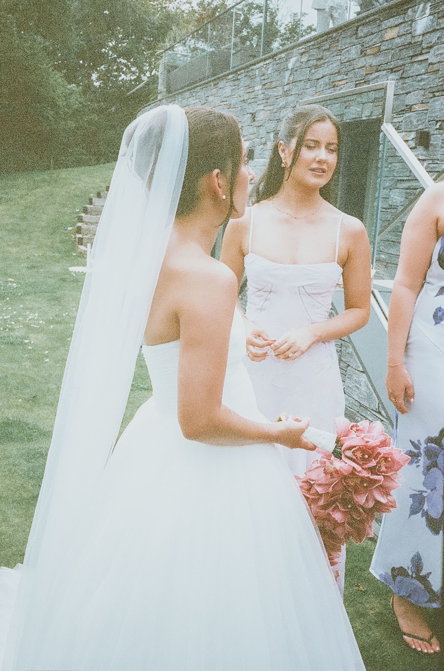 A bride in a white wedding gown with veil stands outdoors with bridesmaids in pink dresses at a stone wall venue.