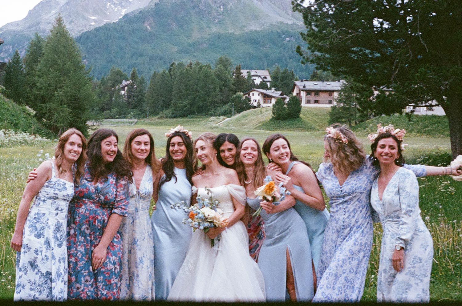 A row of wedding party members in light blue dresses pose together outdoors with mountains in the background.
