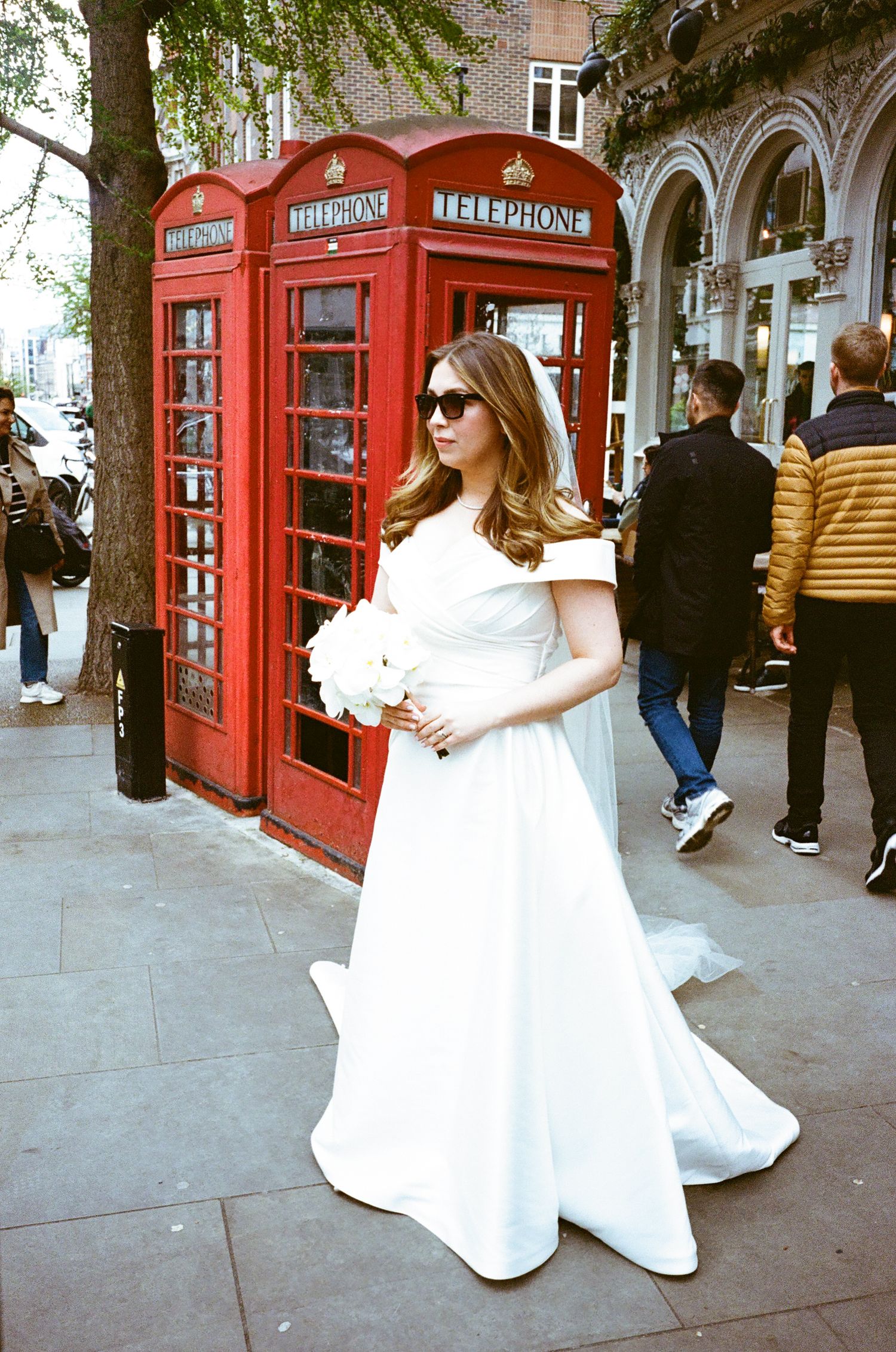 A person in a flowing white dress poses playfully near iconic red British telephone booths on a city street.