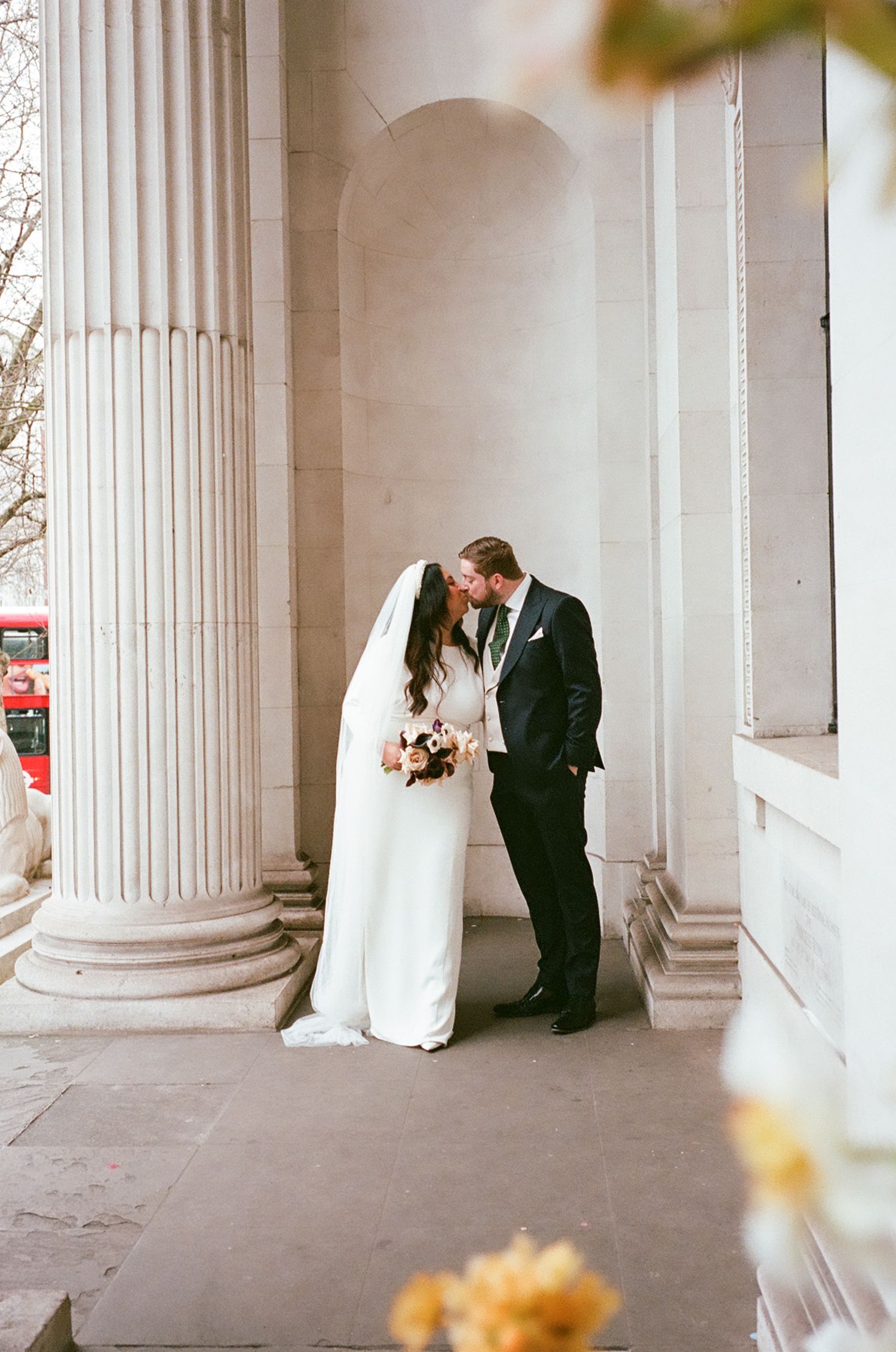 A couple shares an intimate moment between elegant white columns in a classical architectural setting during their wedding.