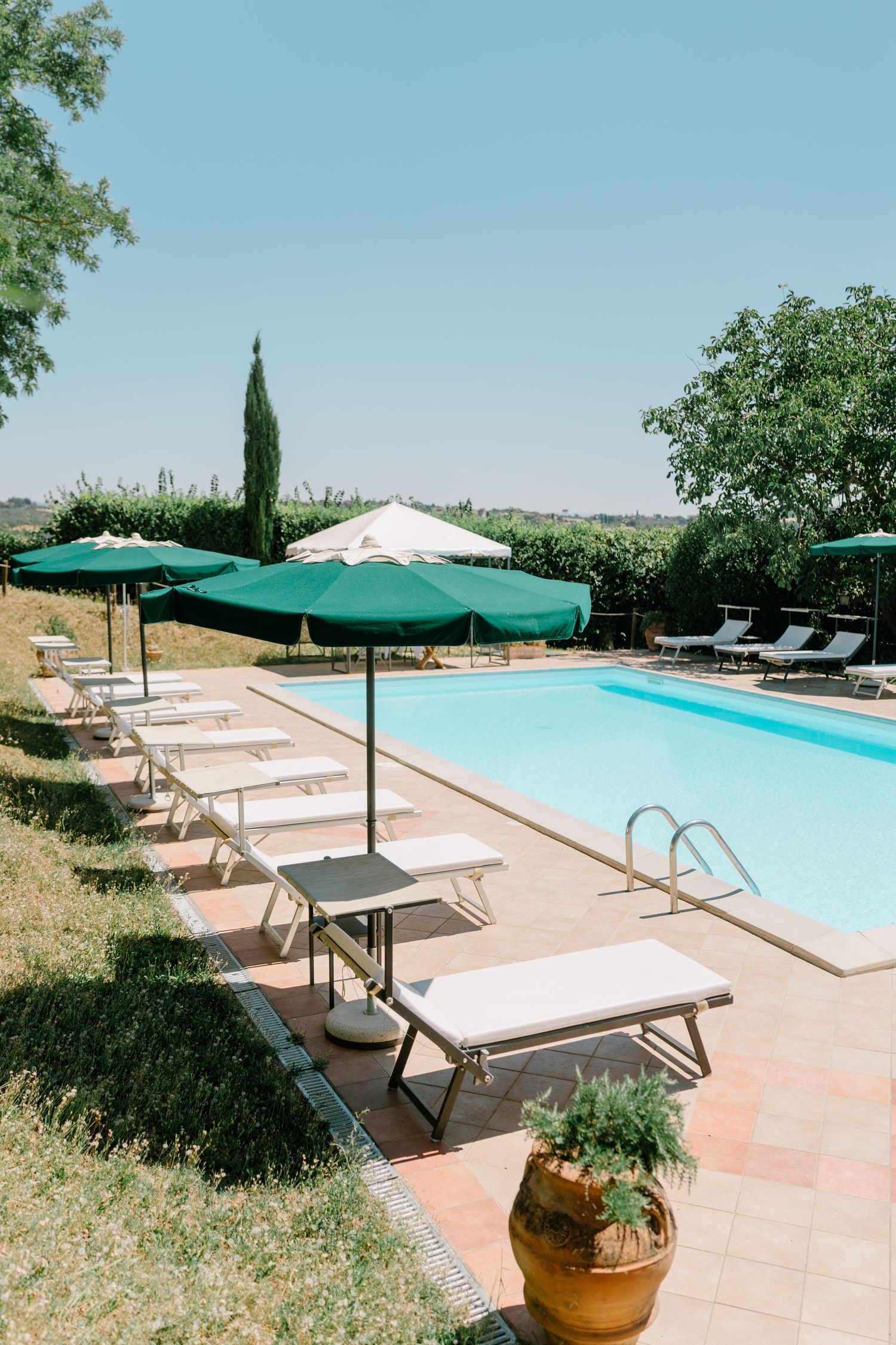 Luxury poolside lounge area with sun beds and green umbrellas overlooking a scenic countryside landscape.