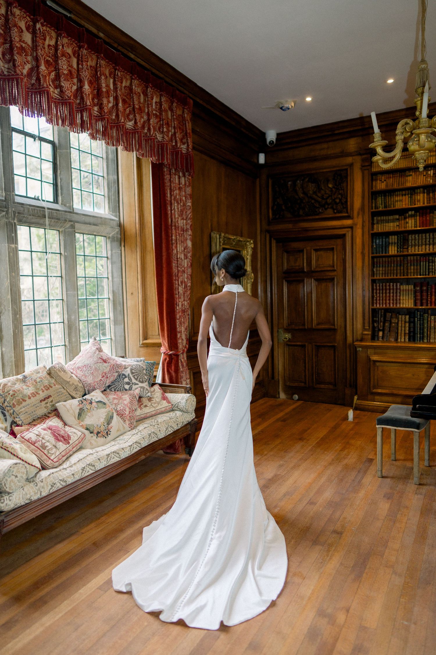 A bride in a white wedding dress poses in a wood-paneled library with vintage furniture and red curtains.