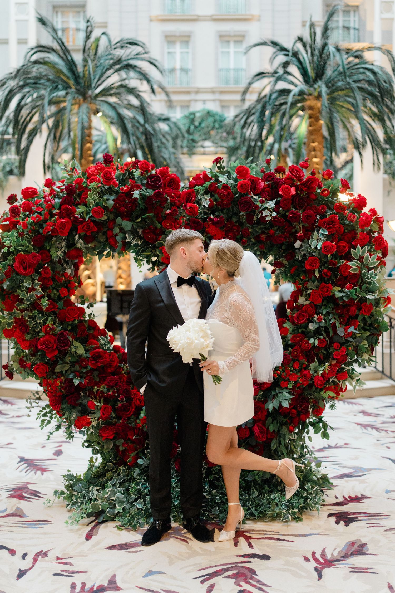 Wedding couple poses by stunning heart-shaped red rose arrangements with palm trees.