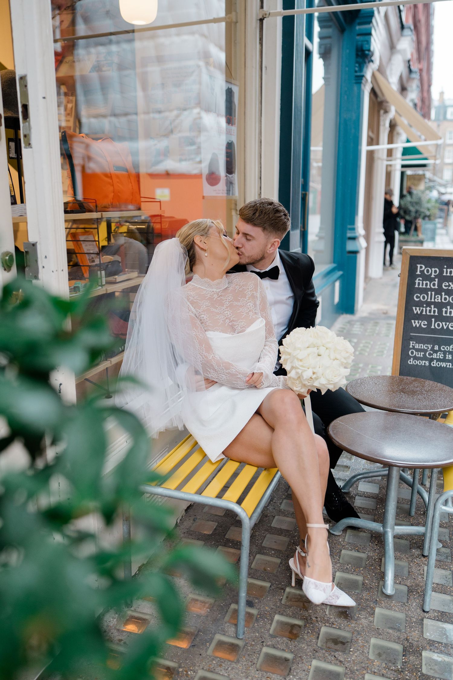 A bride in a short dress sits at an outdoor cafe table in an urban setting.