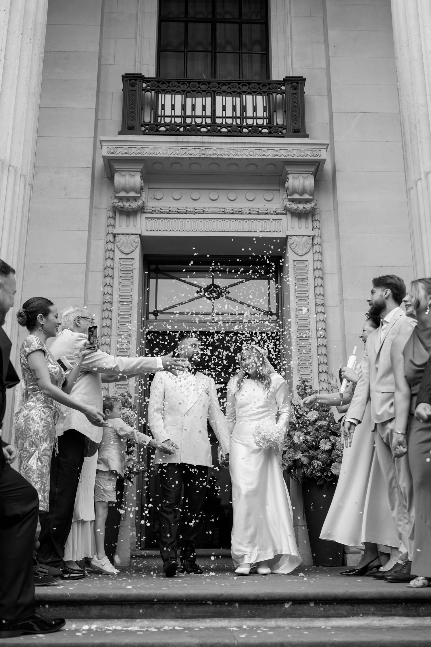 Black and white wedding sequence shows guests throwing confetti at newlyweds outside a grand building entrance.