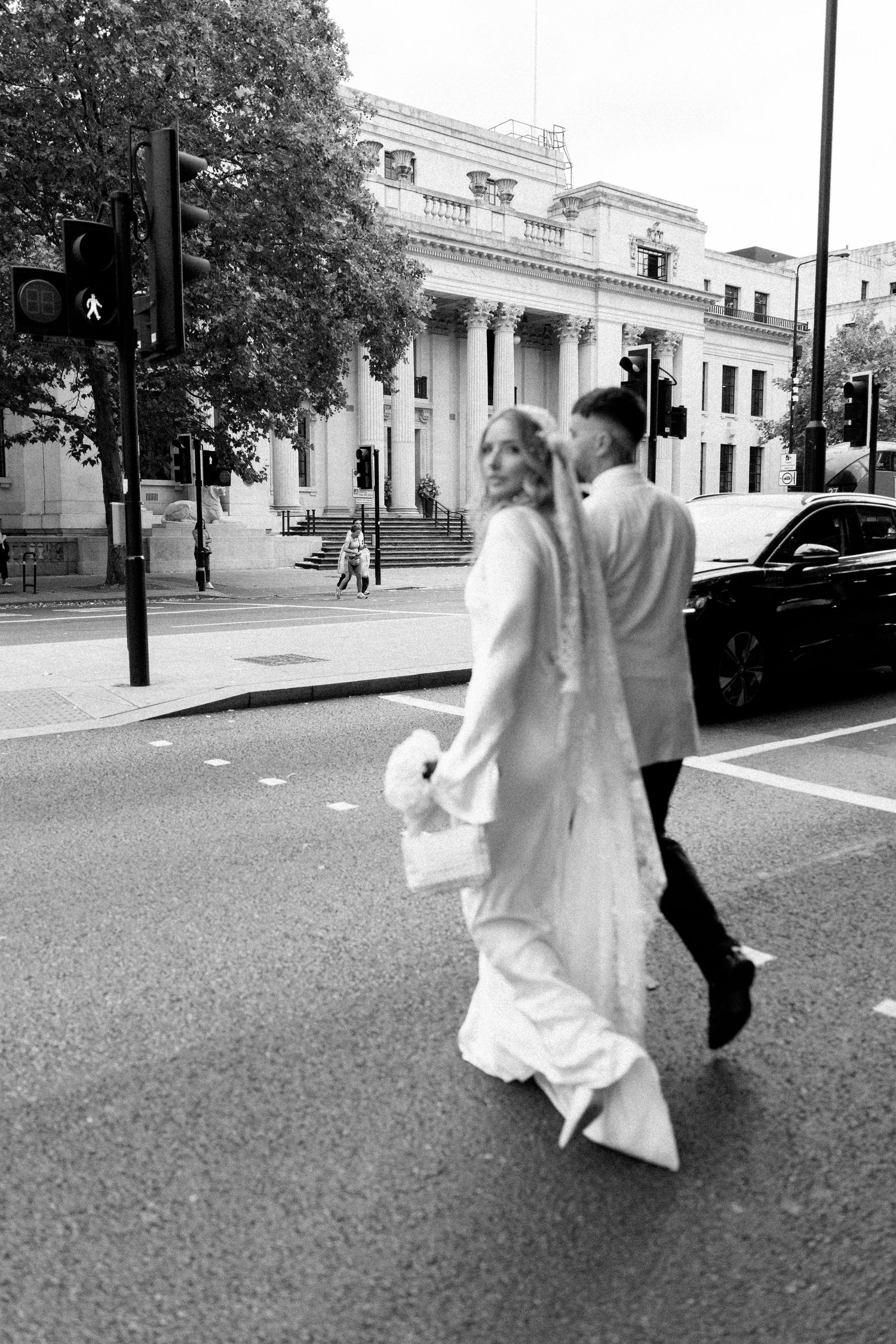 Newlyweds walking away from camera on city street with historic building in background.