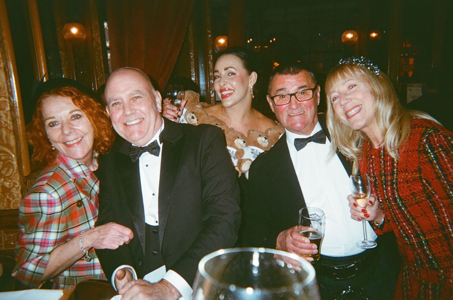 A group of friends in formal attire and casual clothing smile together at an evening social gathering.