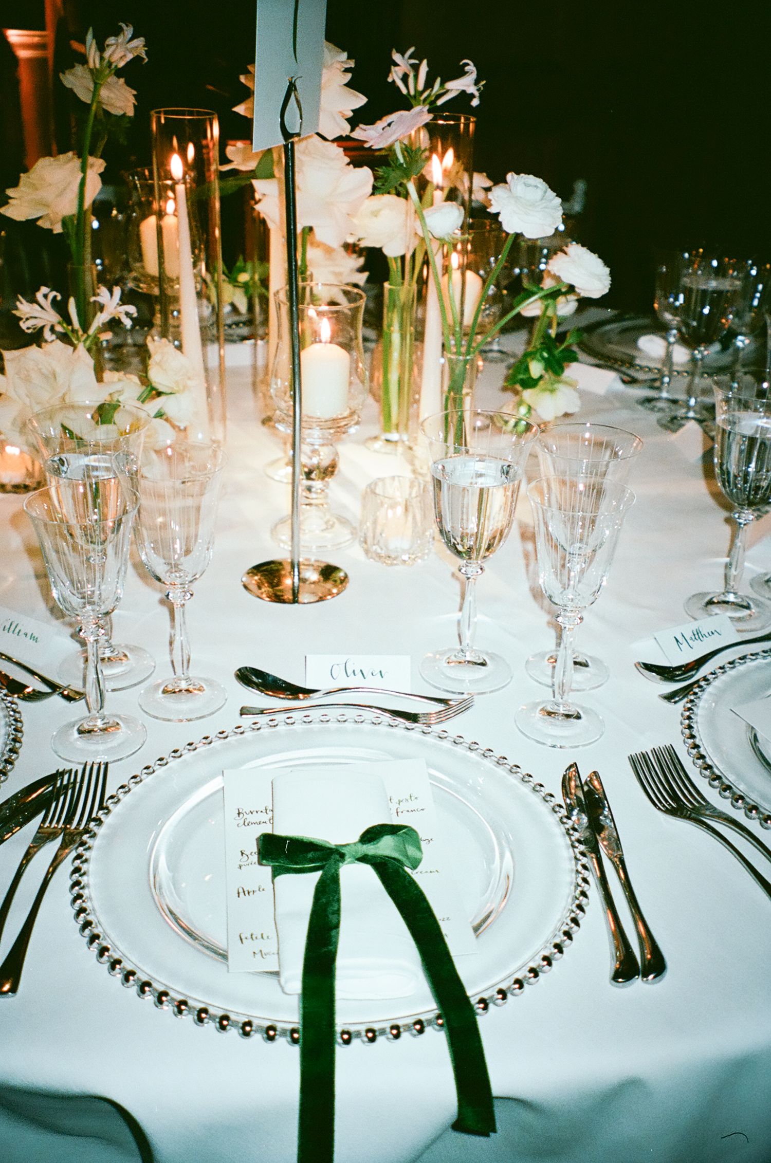 Formal table setting with crystal glasses, white flowers, and green ribbon accents on beaded charger plates.