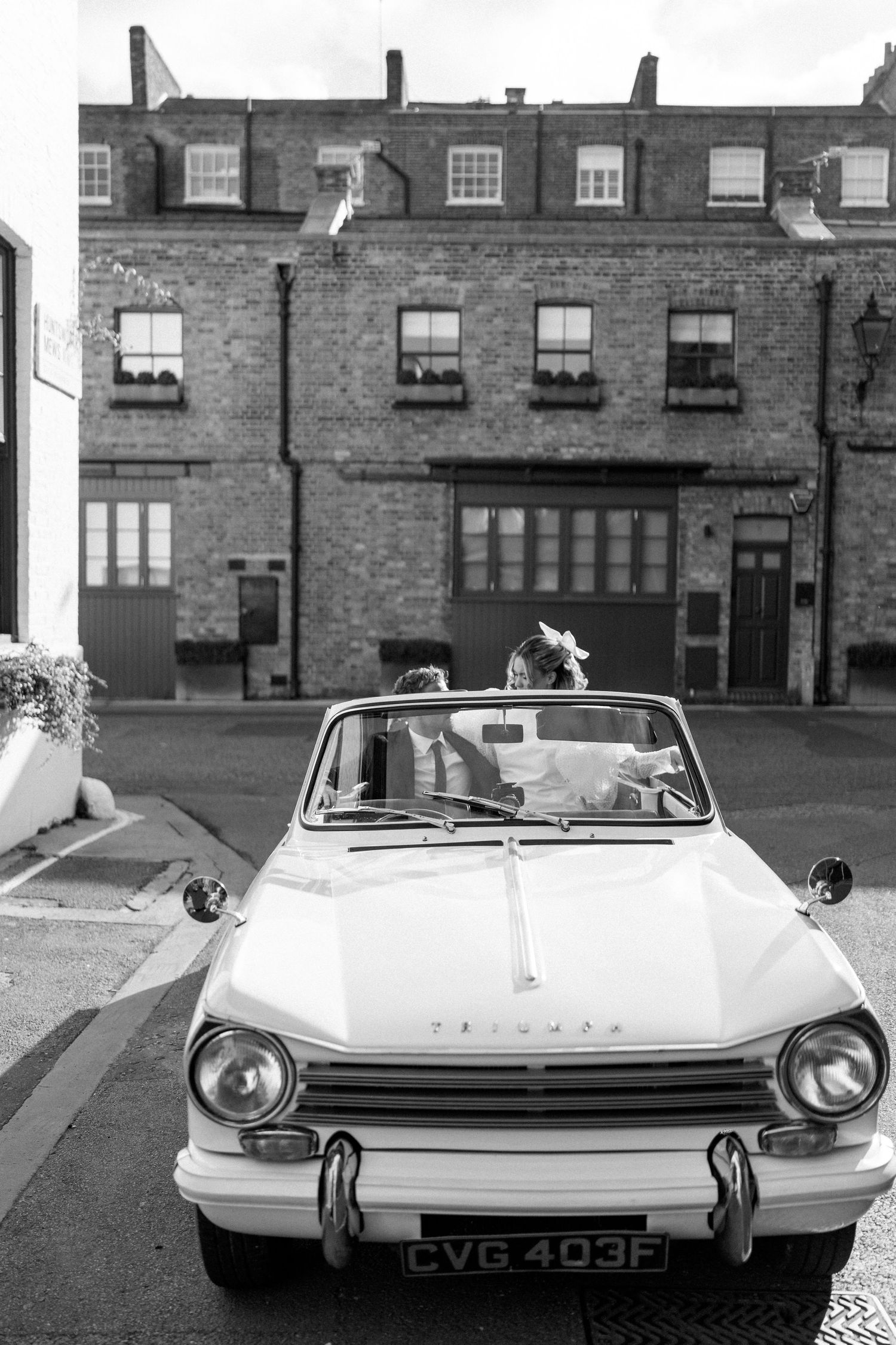 Classic white Triumph car parked in front of brick building in black and white photography.