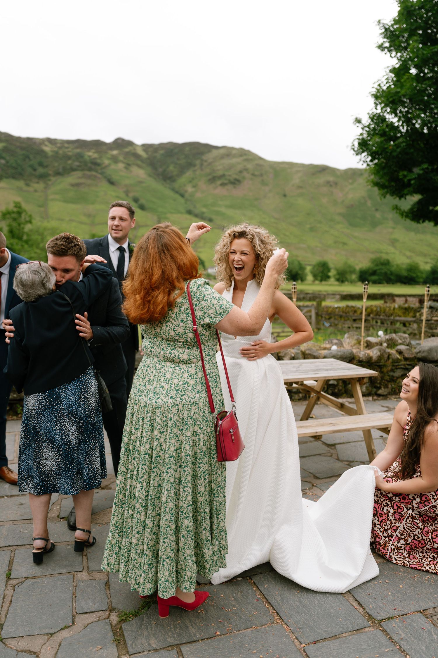 A group of wedding guests laughing and enjoying themselves on a stone patio with green hills in the background.