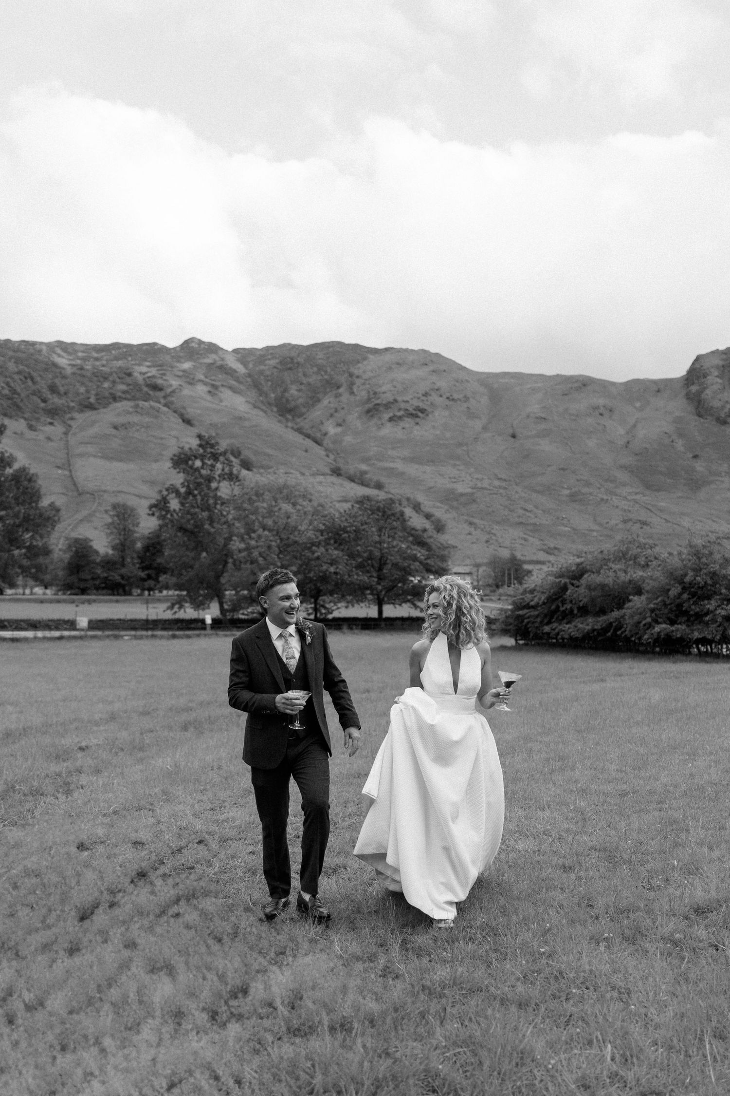 Wedding couple walks playfully across a grassy field with mountains in the background.