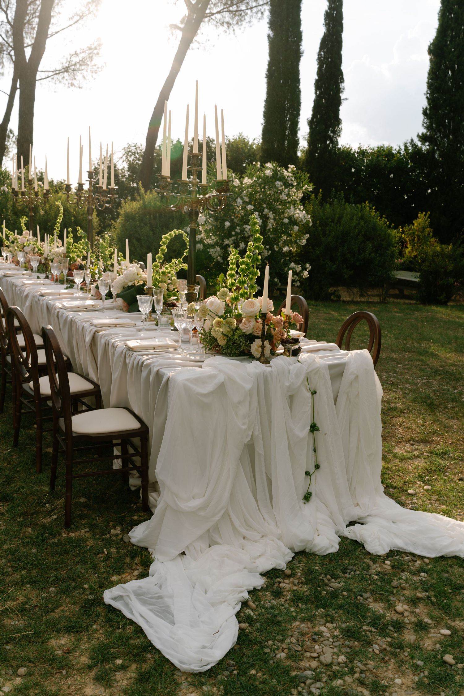Long wedding reception table with white linens and tall candles in an outdoor garden setting.