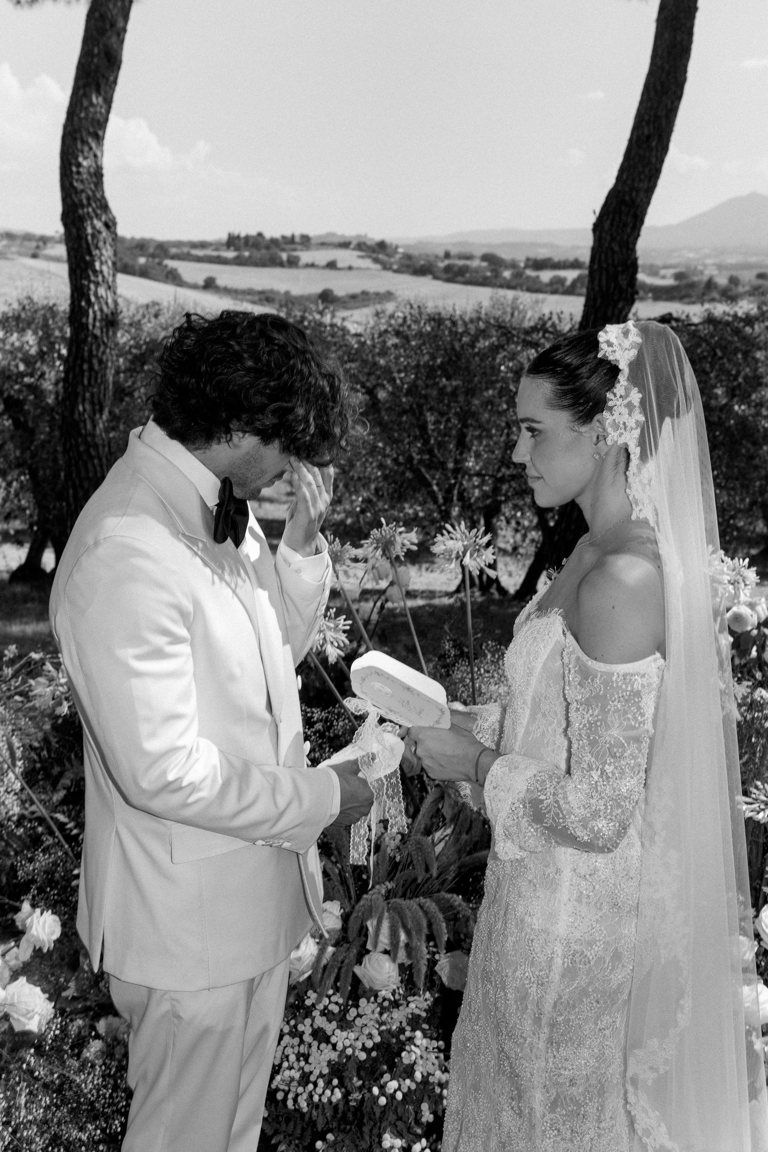 Outdoor wedding ceremony under trees with couple exchanging vows in white attire.