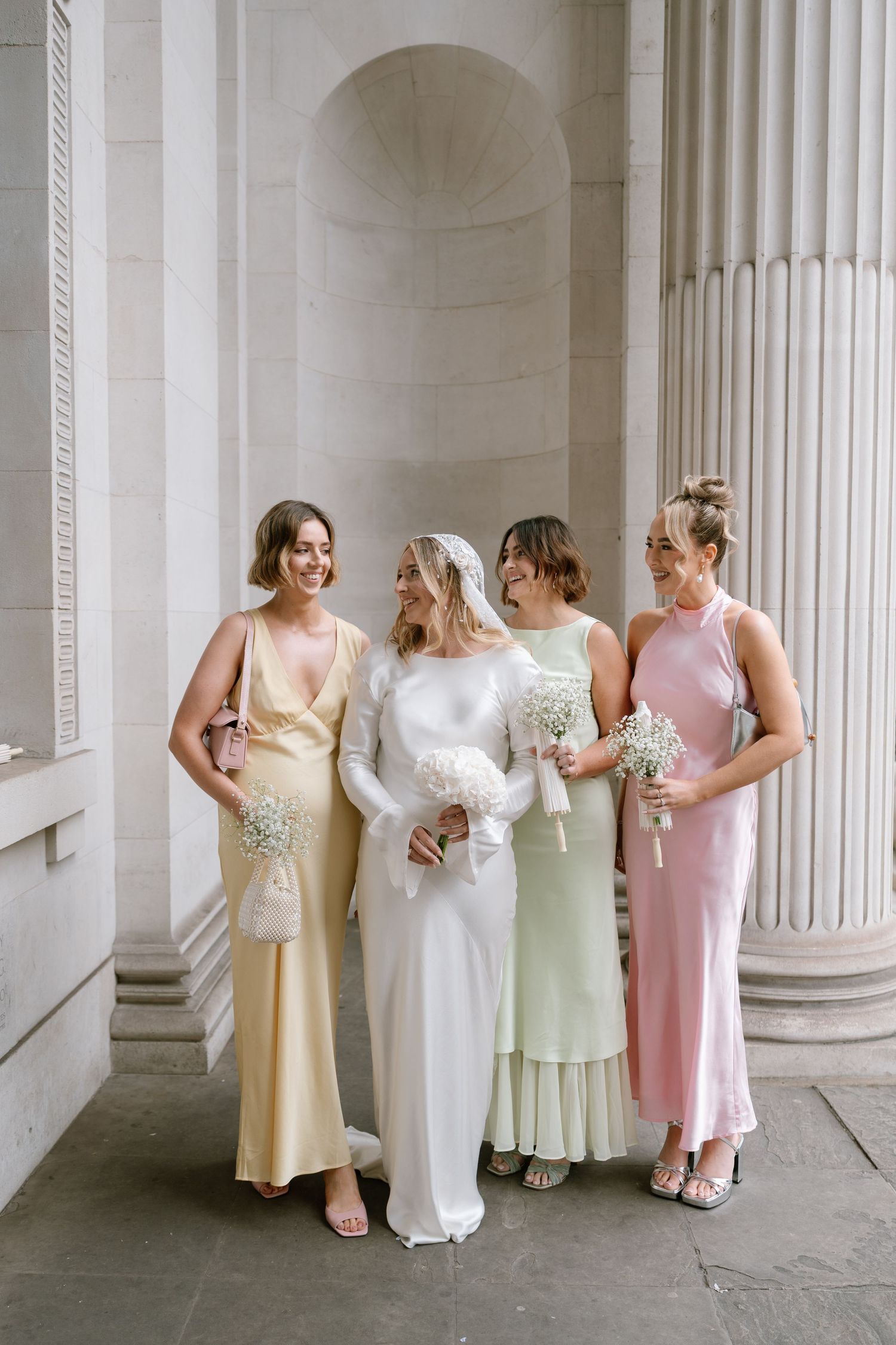 Wedding party in pastel dresses posing together against white columns in an elegant venue.
