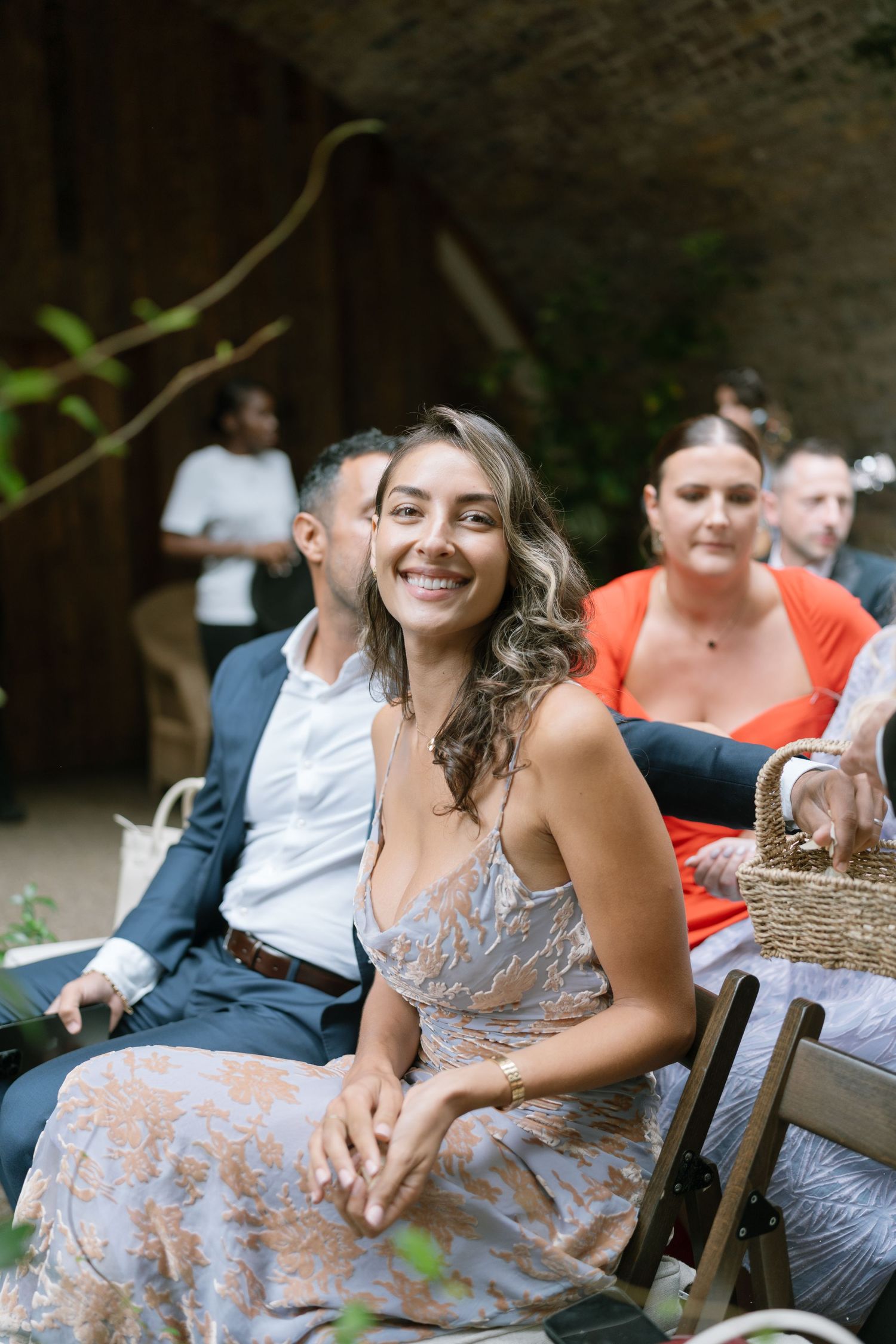 Wedding guest in a floral dress sitting on a wooden bench during an outdoor ceremony.