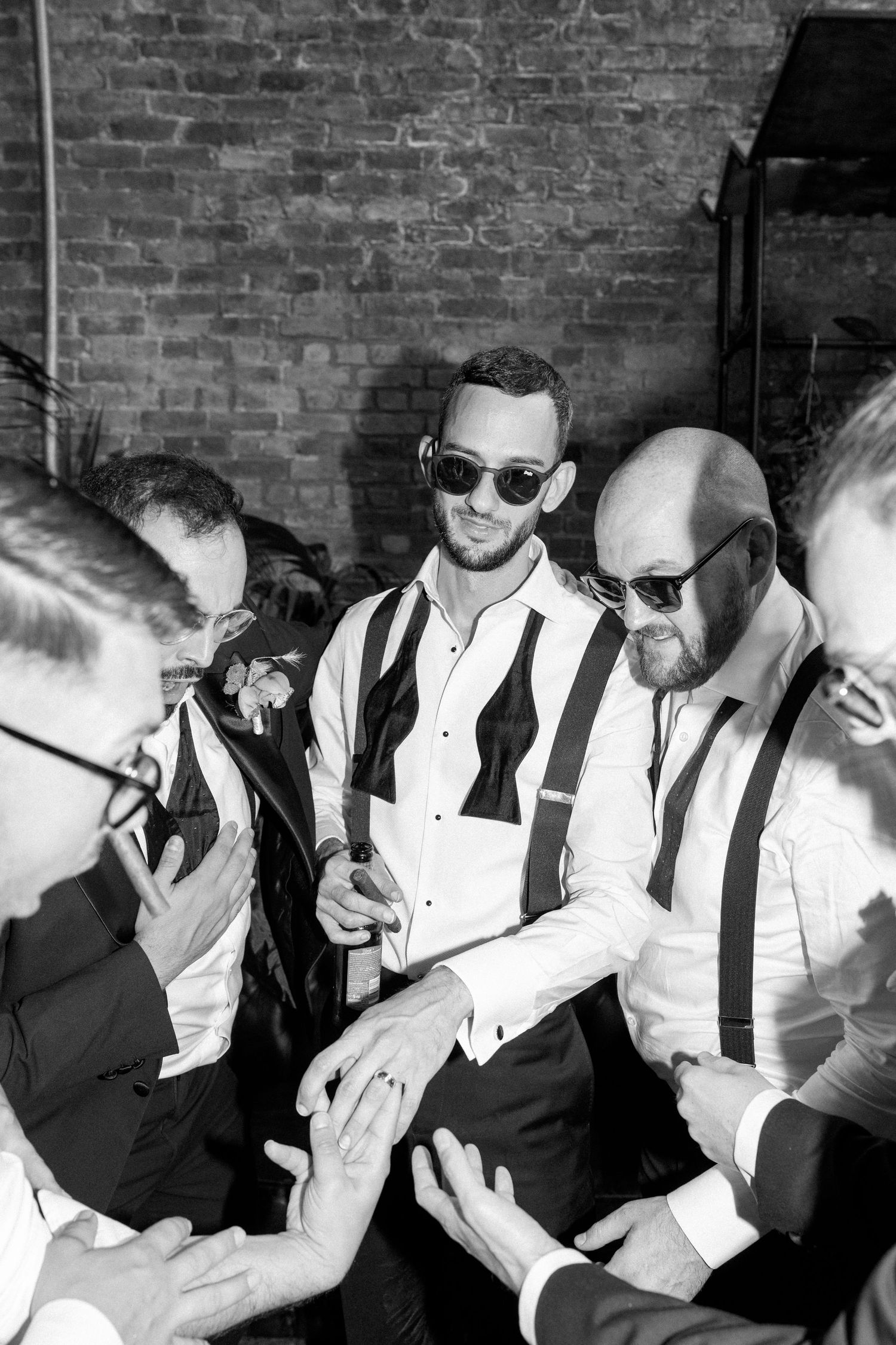 Groomsmen in formal attire and sunglasses laugh together at wedding reception.