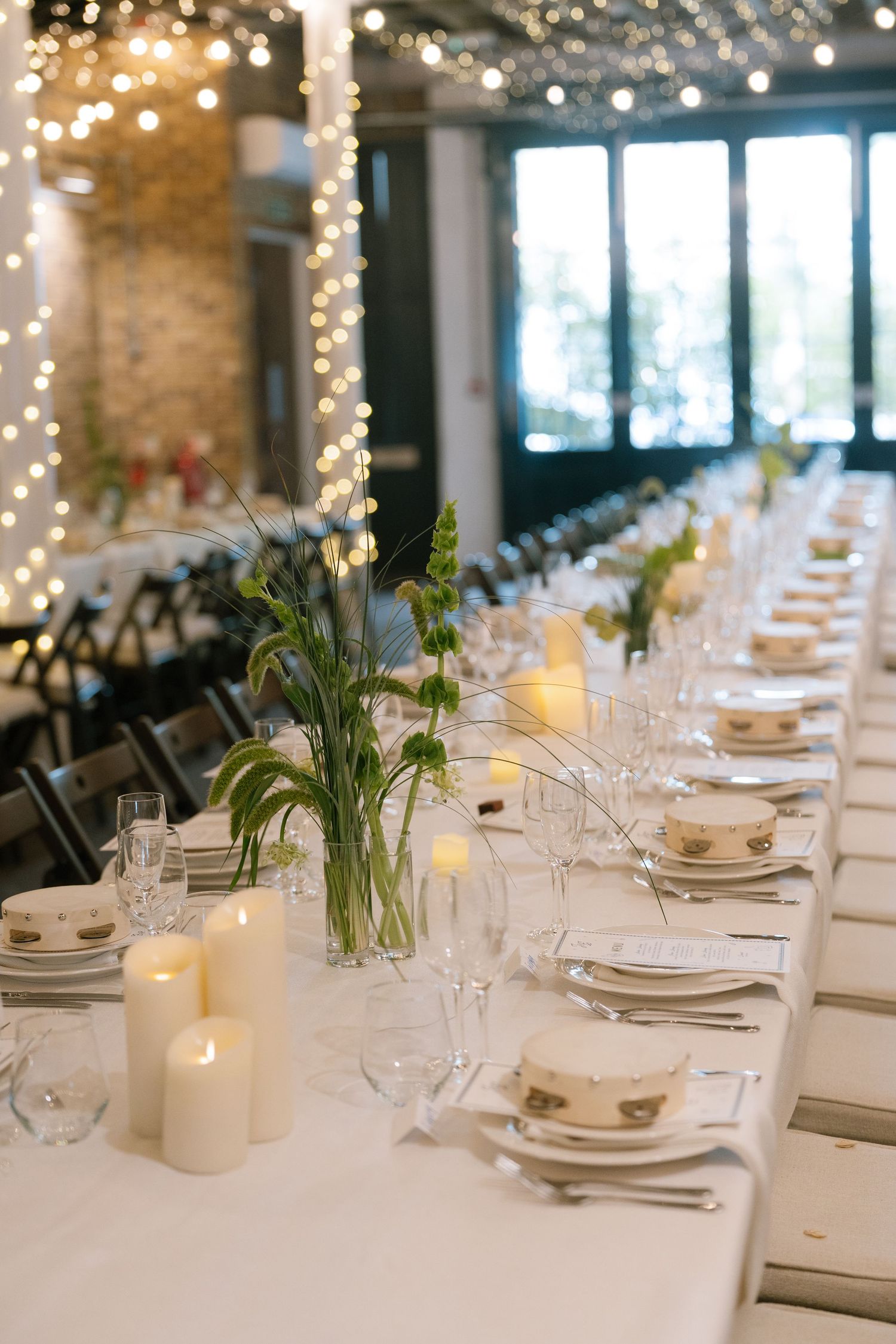 Elegant wedding reception table settings with white linens, candles and string lights in an industrial venue.