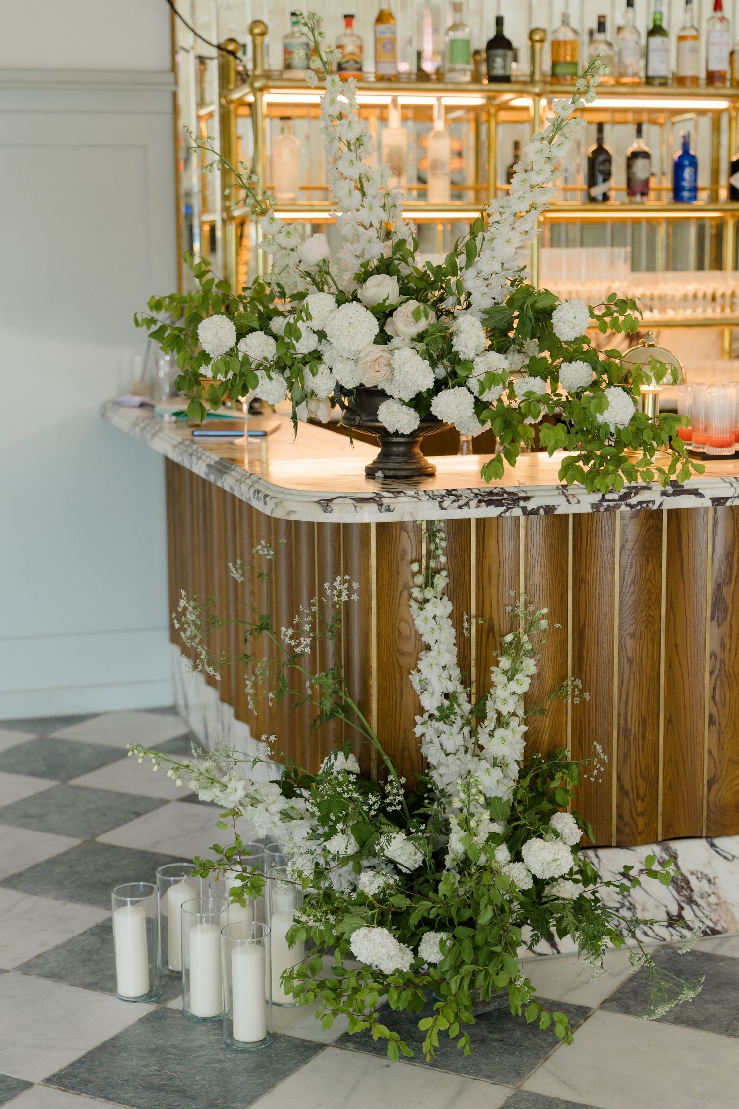 Elegant wedding bar decorated with lush white floral arrangements, greenery, and pillar candles on checkered floor.