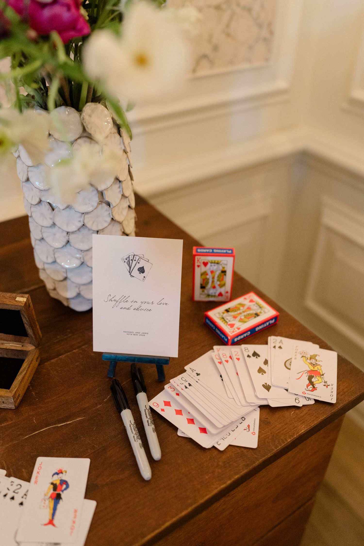 Playing card guest book station with Sharpie markers, card deck, and sign on wooden table with floral arrangement.