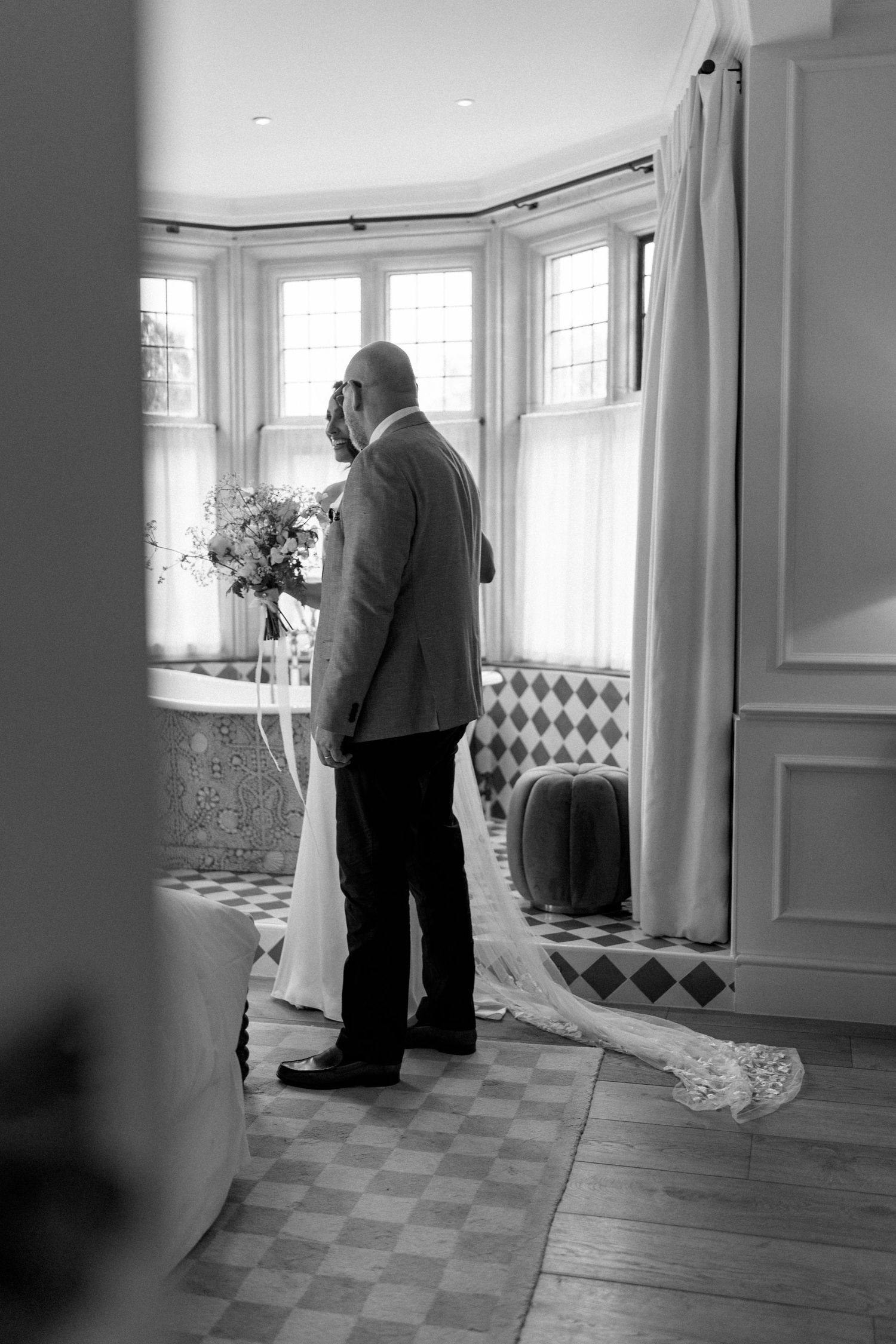 Black and white photo of a man holding a bridal bouquet standing by bay windows in a checkered bathroom.