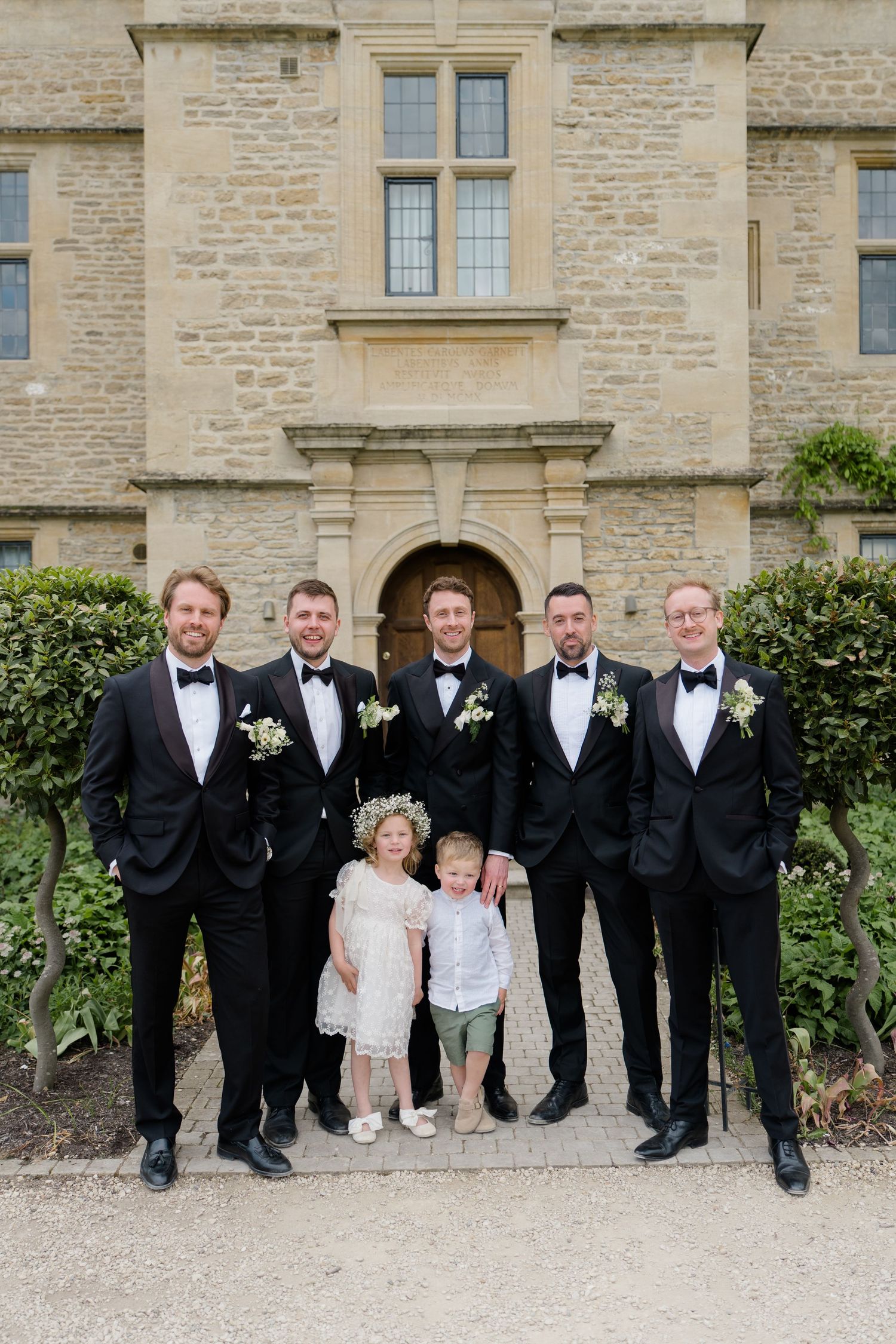 Groomsmen in black tuxedos posing with two children outside stone manor wedding venue.