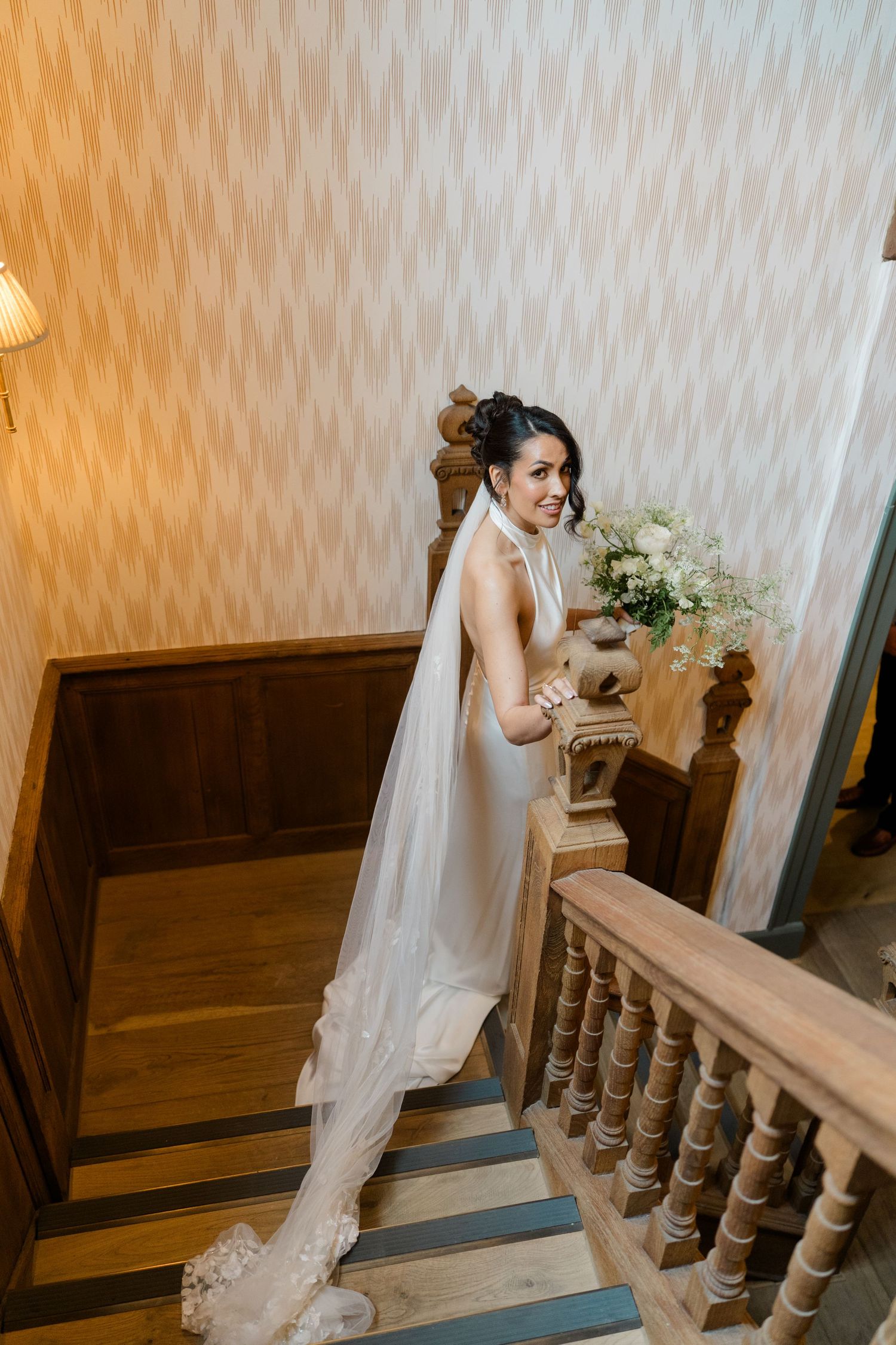 Bride in elegant backless wedding gown with long veil descending staircase holding bouquet.