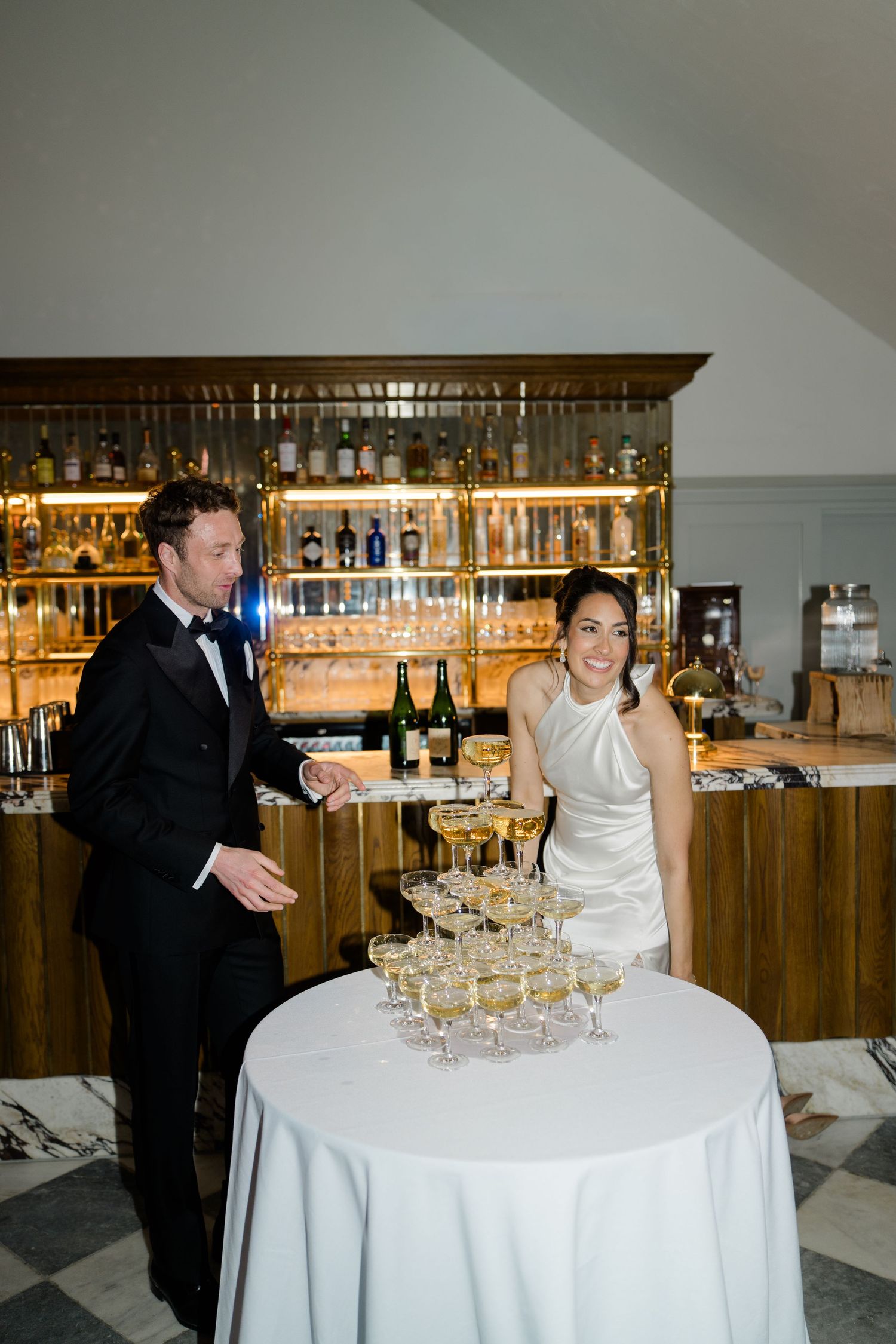 Bride and groom smile beside champagne tower at elegant bar during wedding reception.