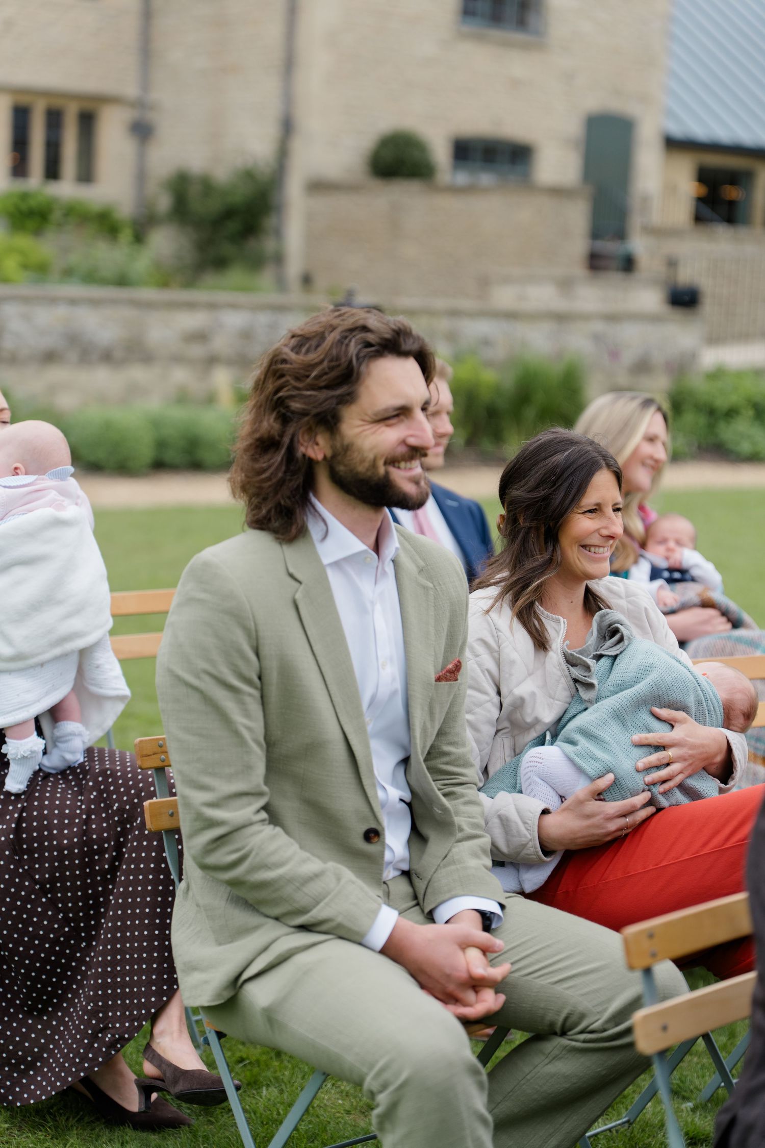 Smiling wedding guests seated outdoors on wooden chairs, man in sage green suit beside woman holding sleeping baby.