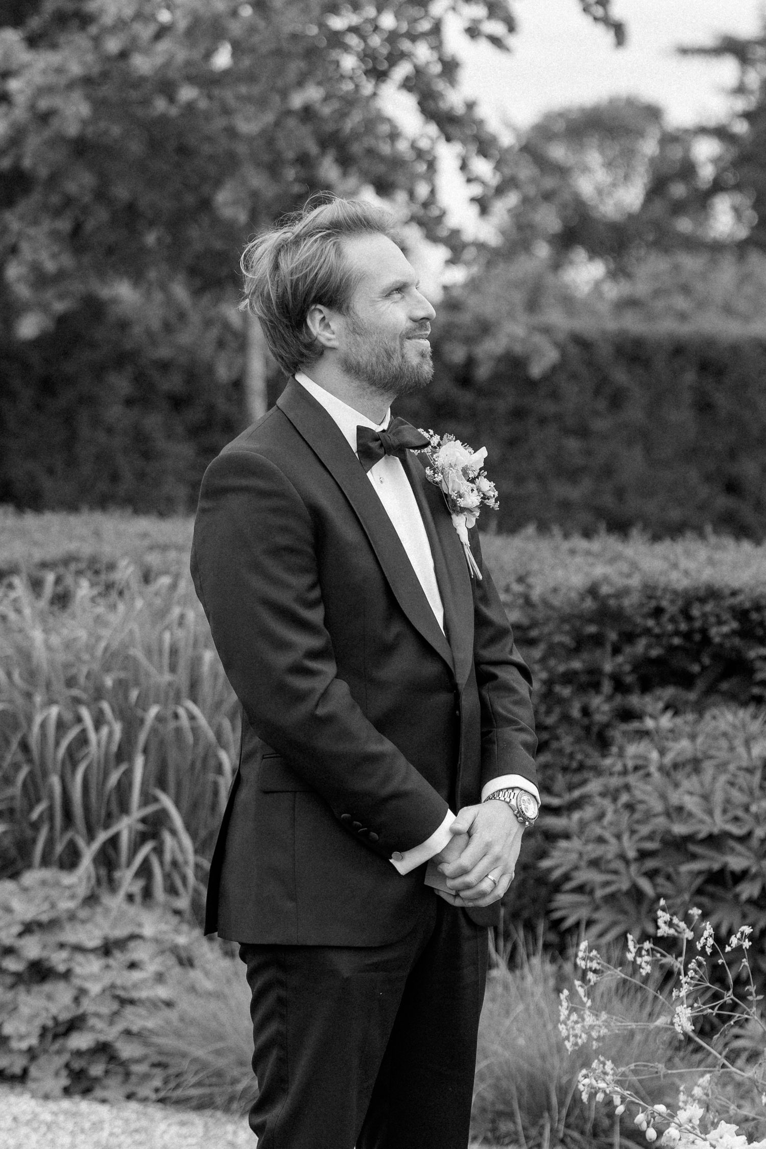 Black and white portrait of handsome groom in tuxedo with floral boutonniere standing in garden.