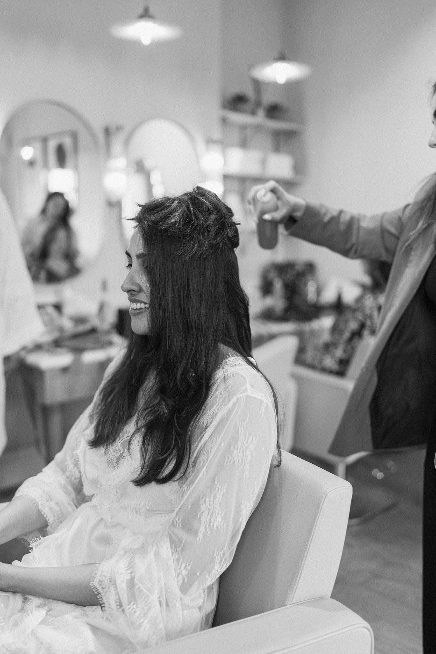 Black and white photo of smiling bride getting hair styled in white lace robe at salon.