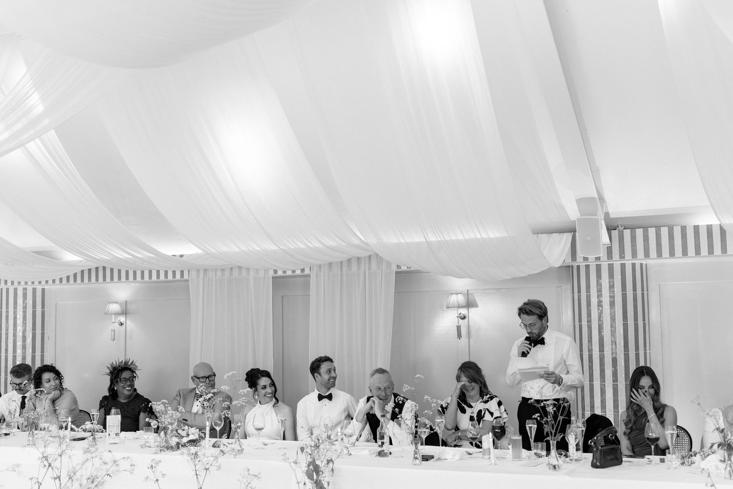 Wedding party seated at head table during reception speech in elegant white draped venue.