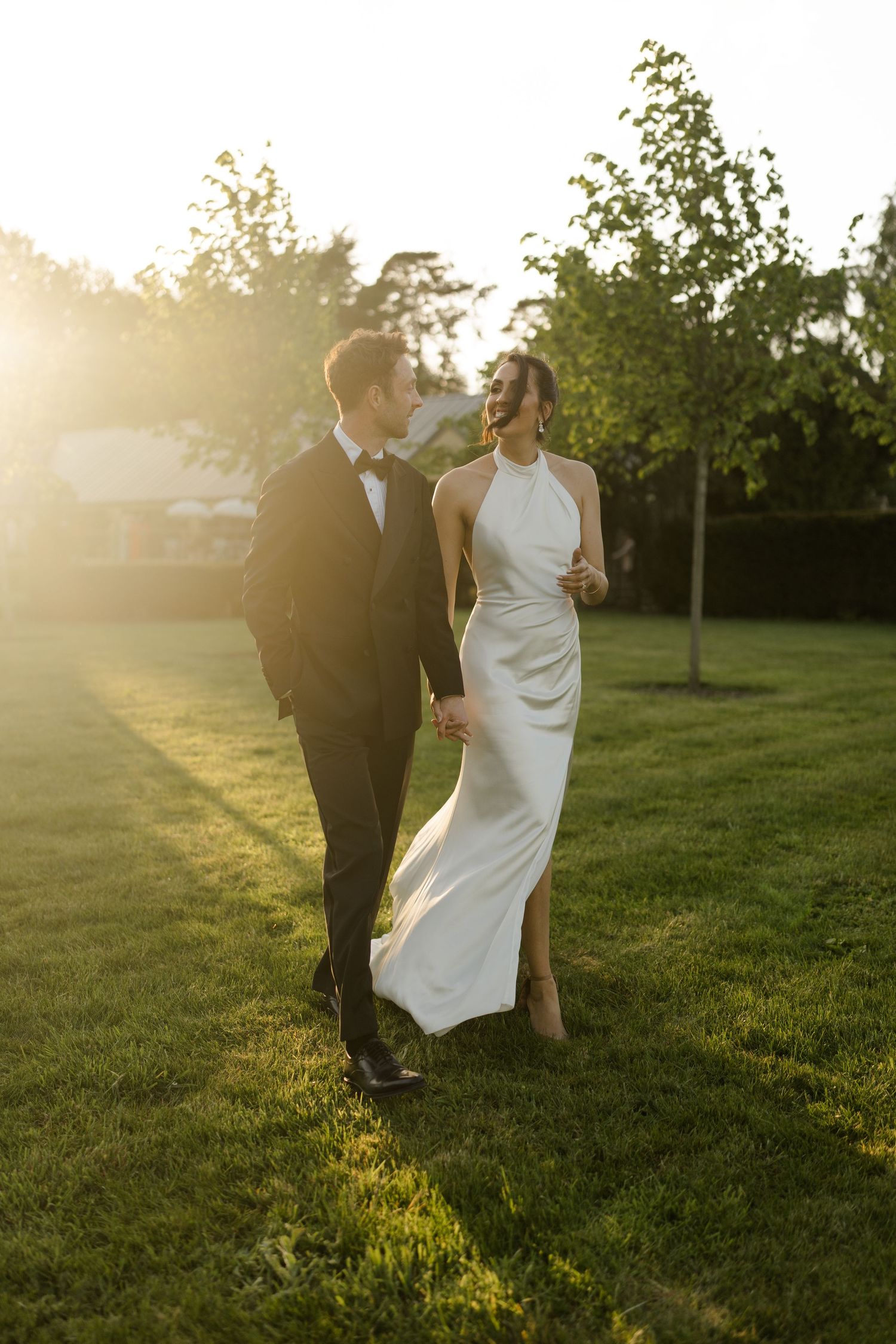 Bride and groom walking hand in hand through golden sunset light on lush green lawn.