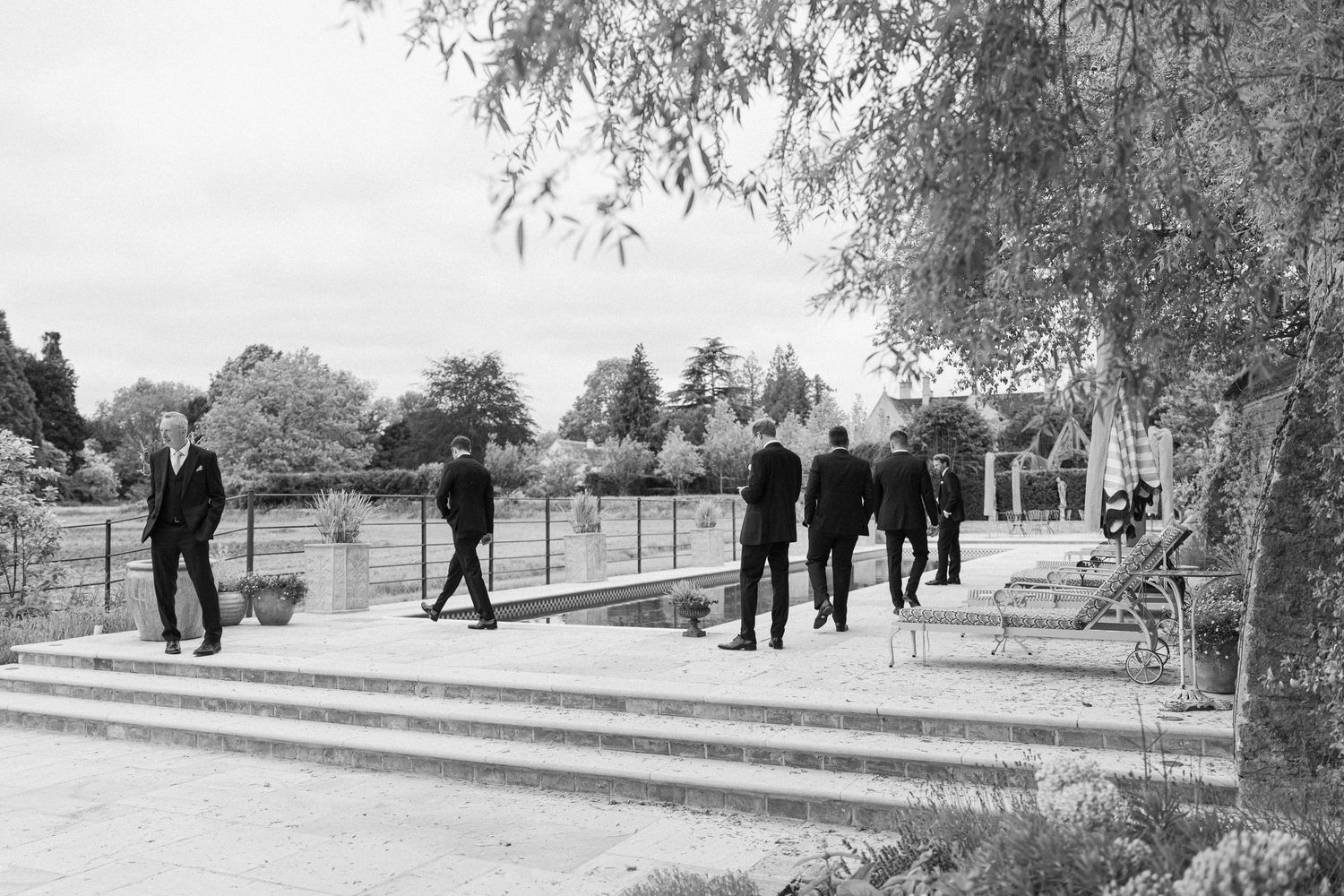 Black and white photo of groomsmen walking on stone terrace surrounded by lush garden landscape.