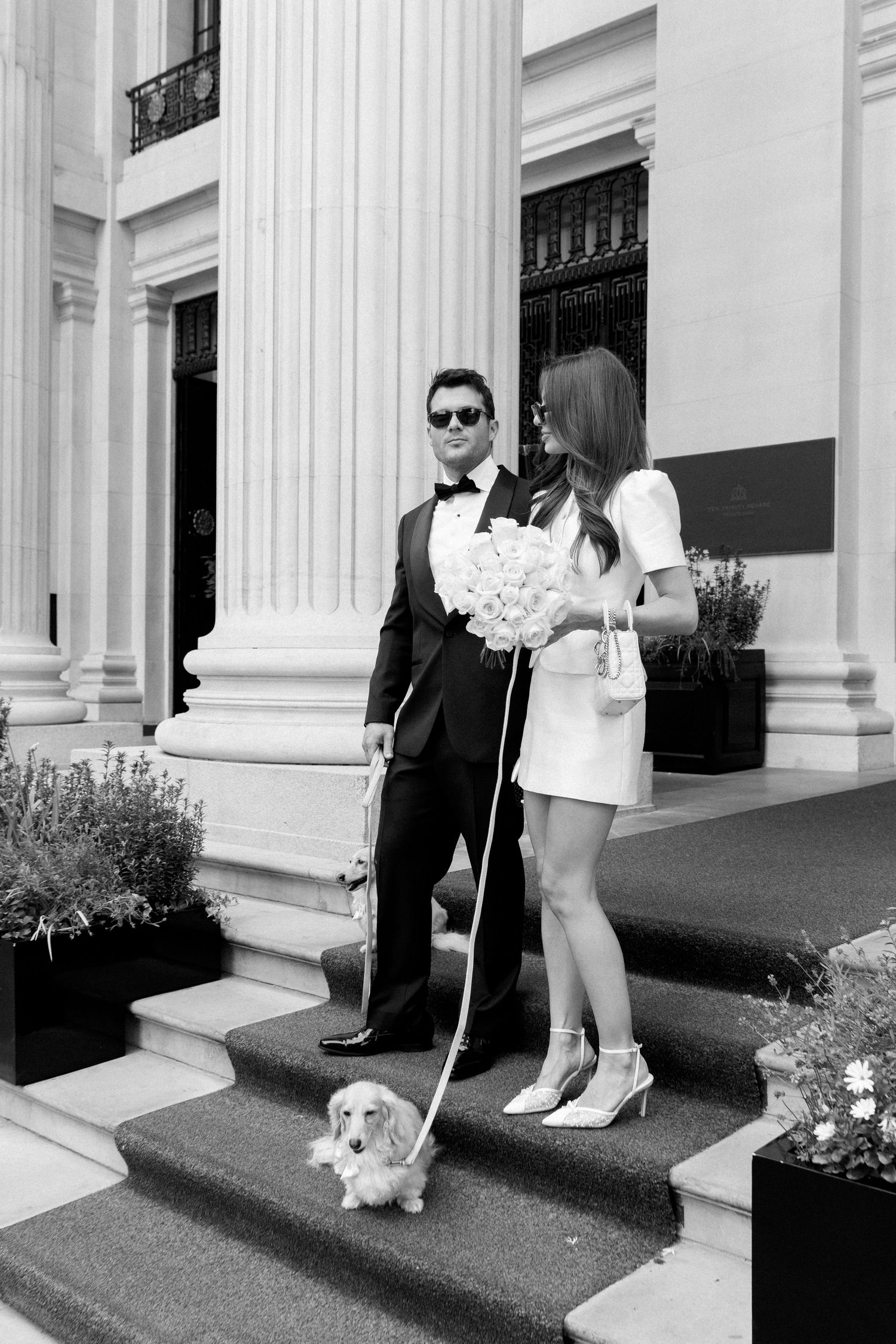 Bride and groom in formal wedding attire pose on hotel steps with long-haired dachshund on leash.