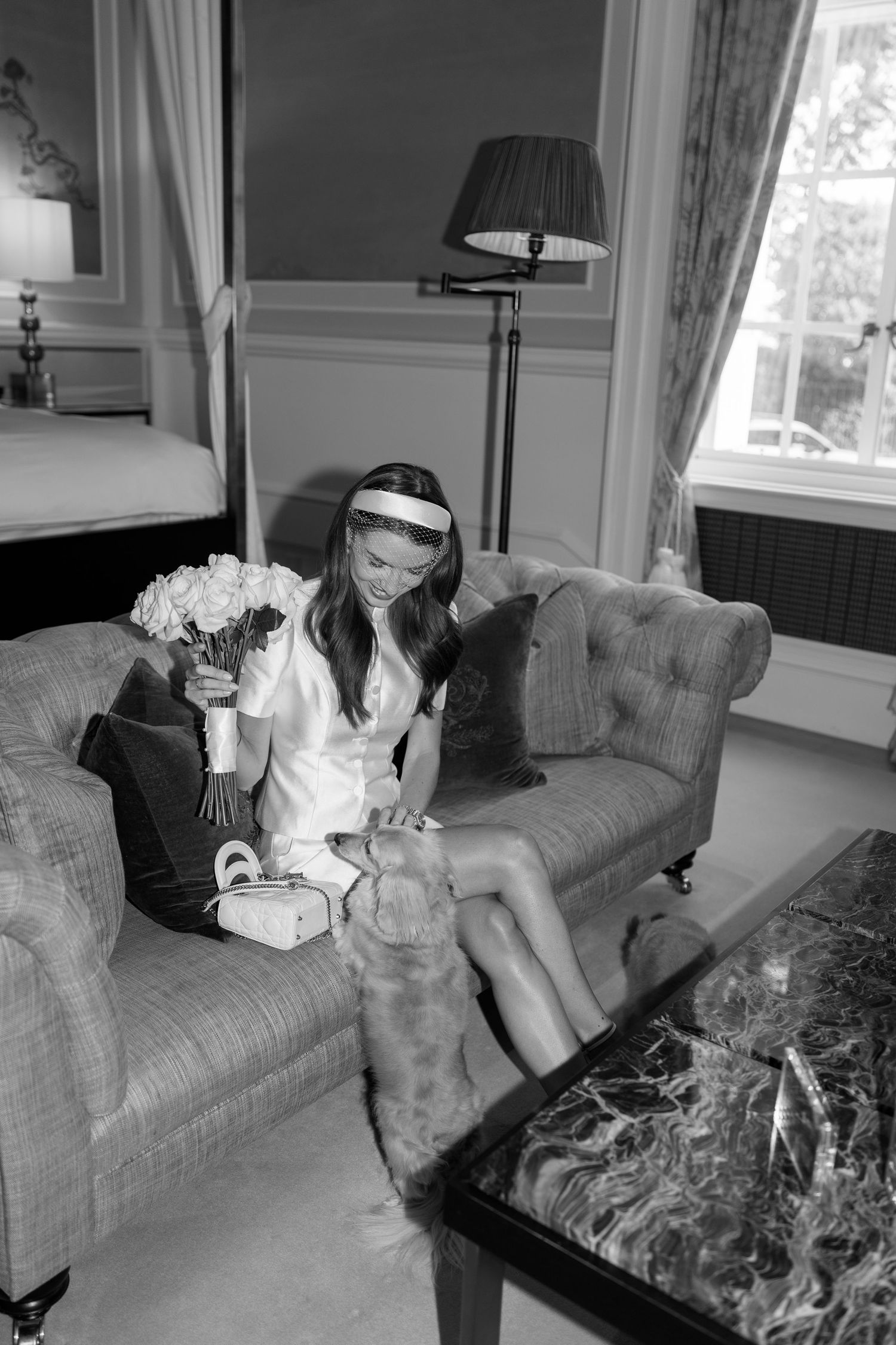 Smiling bride in white dress holds bouquet while sitting on sofa with small dog in elegant room.