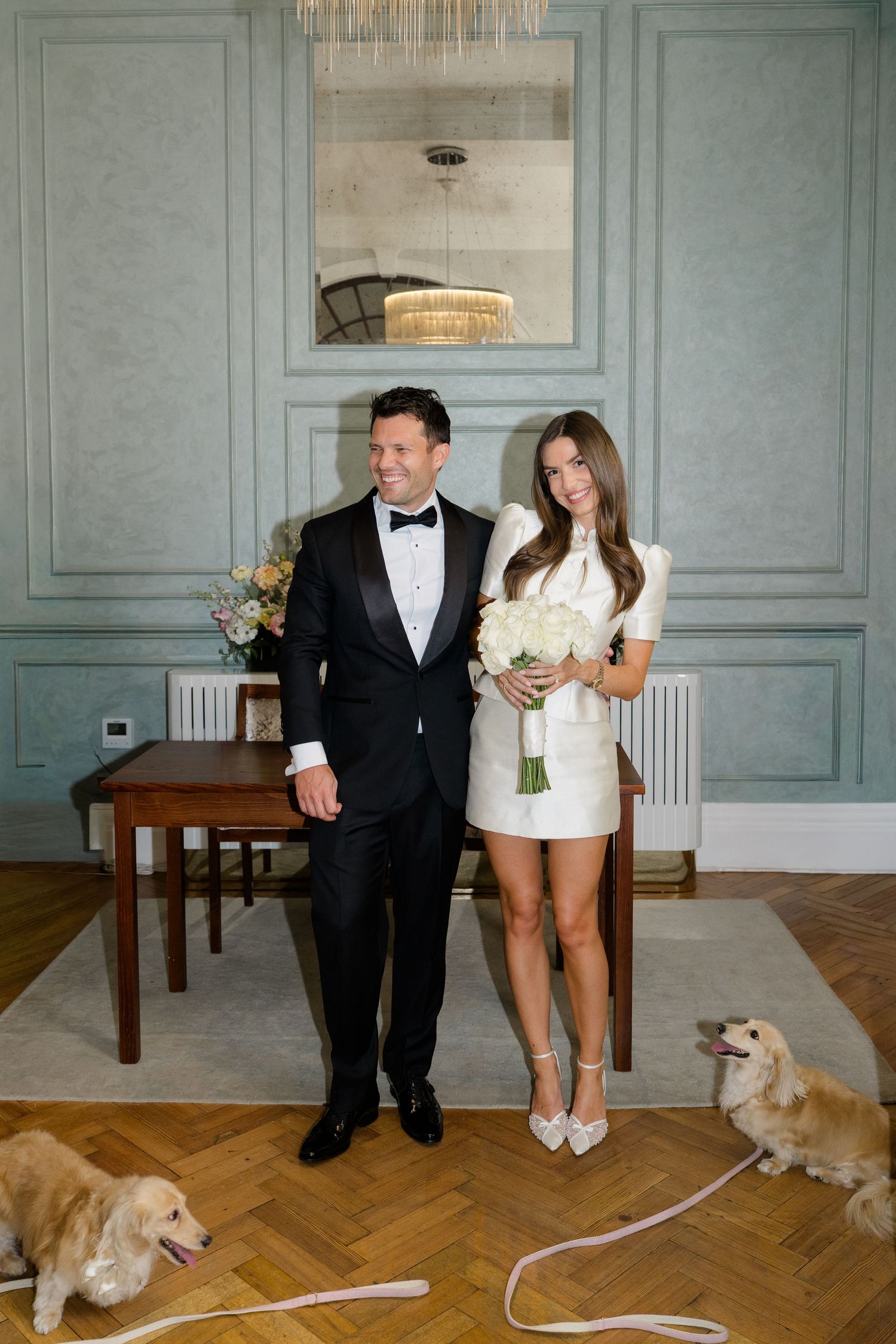 Bride in white mini dress and groom in tuxedo stand with two dogs in elegant room.